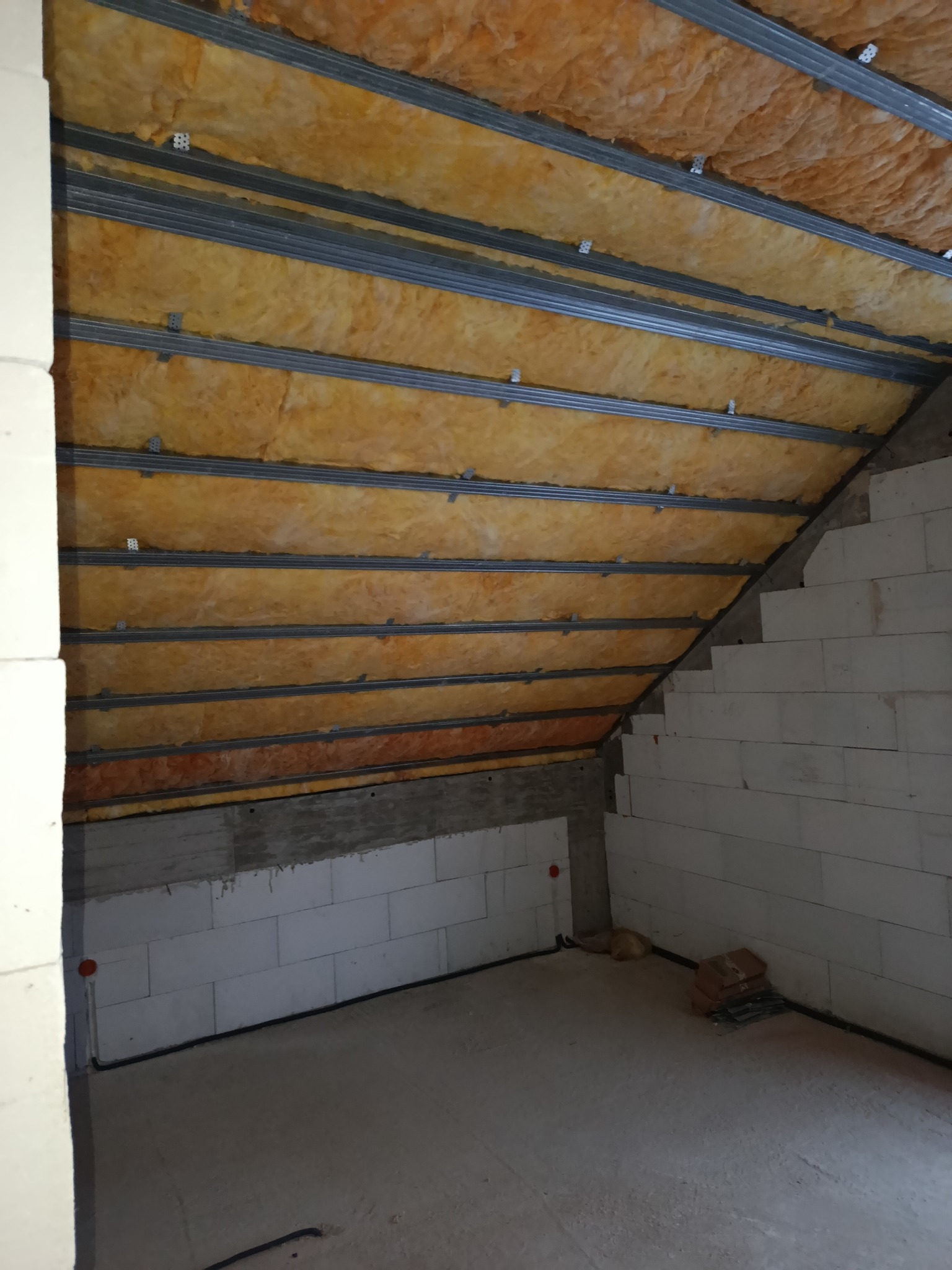 Skośne poddasze z widoczną izolacją z wełny mineralnej między metalowymi profilami, ściany z białych bloczków. Etap prac przed wykończeniem.