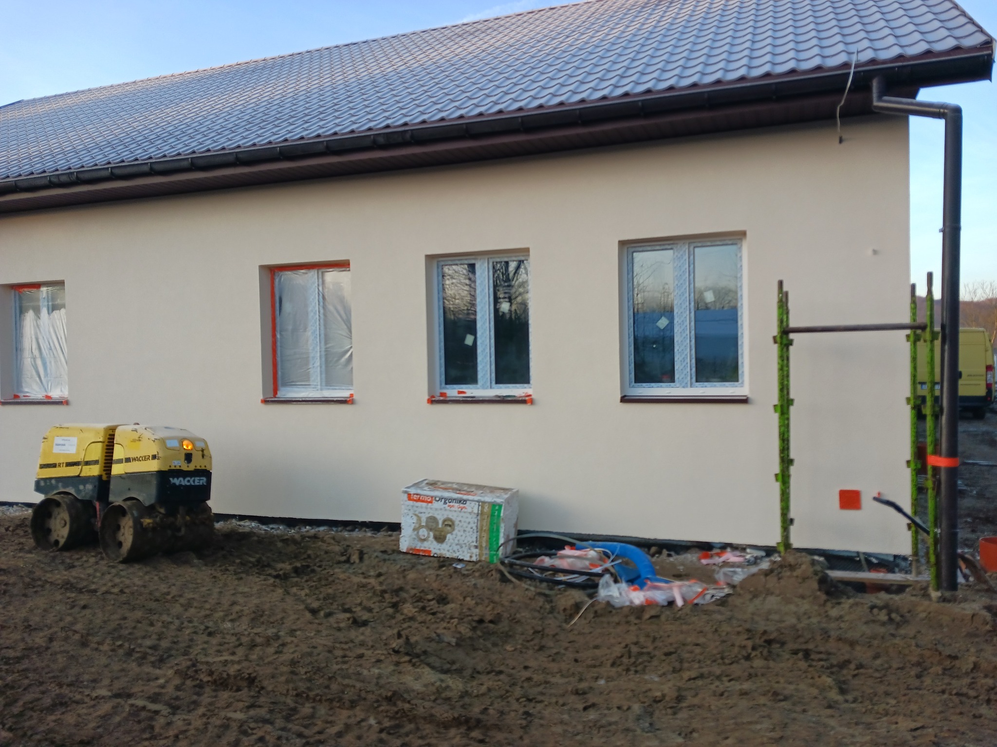 Jasna elewacja domu jednorodzinnego z oknami zabezpieczonymi folią. Na pierwszym planie ubijarka gruntu Wacker i materiały budowlane. Widoczne rusztowanie.