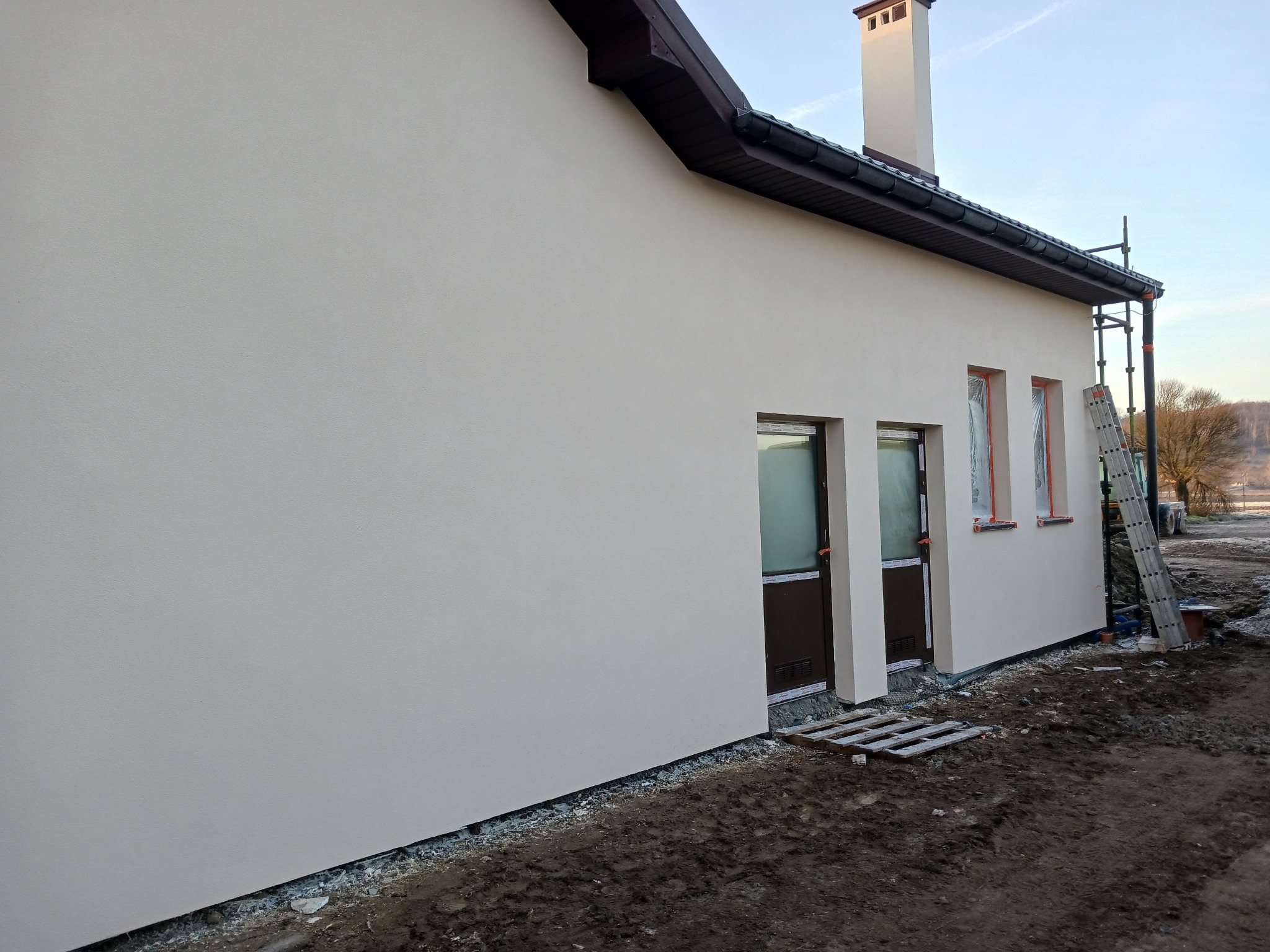 Jasna elewacja domu jednorodzinnego po tynkowaniu, z widocznymi oknami zabezpieczonymi folią i drabiną opartą o ścianę. Świeżo wykończona ściana w neutralnym odcieniu.