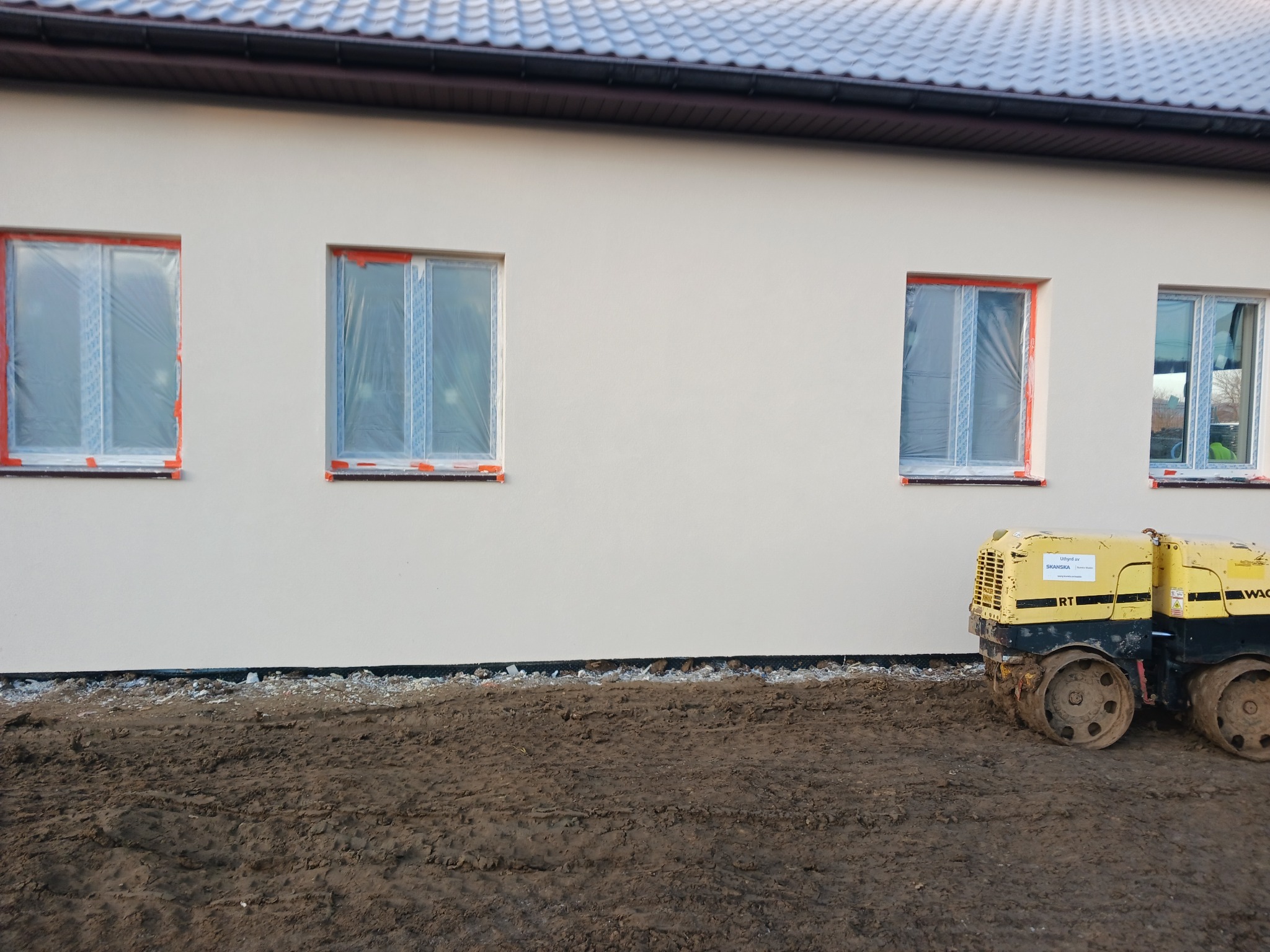 Ściana budynku po tynkowaniu, okna zabezpieczone folią, obok stoi walec do ubijania ziemi. Widoczny fragment dachu z brązową podbitką.