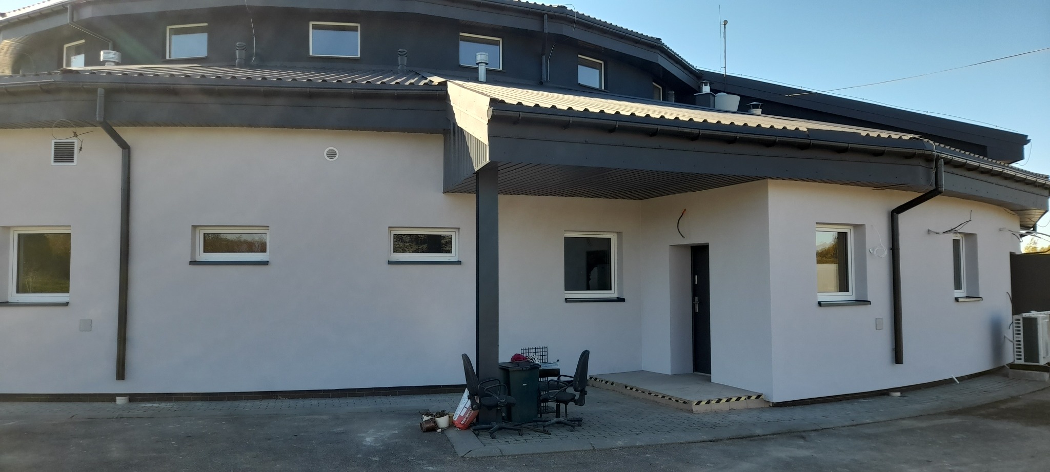 Jasnoszara elewacja budynku z ciemnoszarą stolarką okienną i dachem. Widoczne okna, rynny i fragment otoczenia. Uwagę zwraca nietypowy, zaokrąglony kształt budynku.