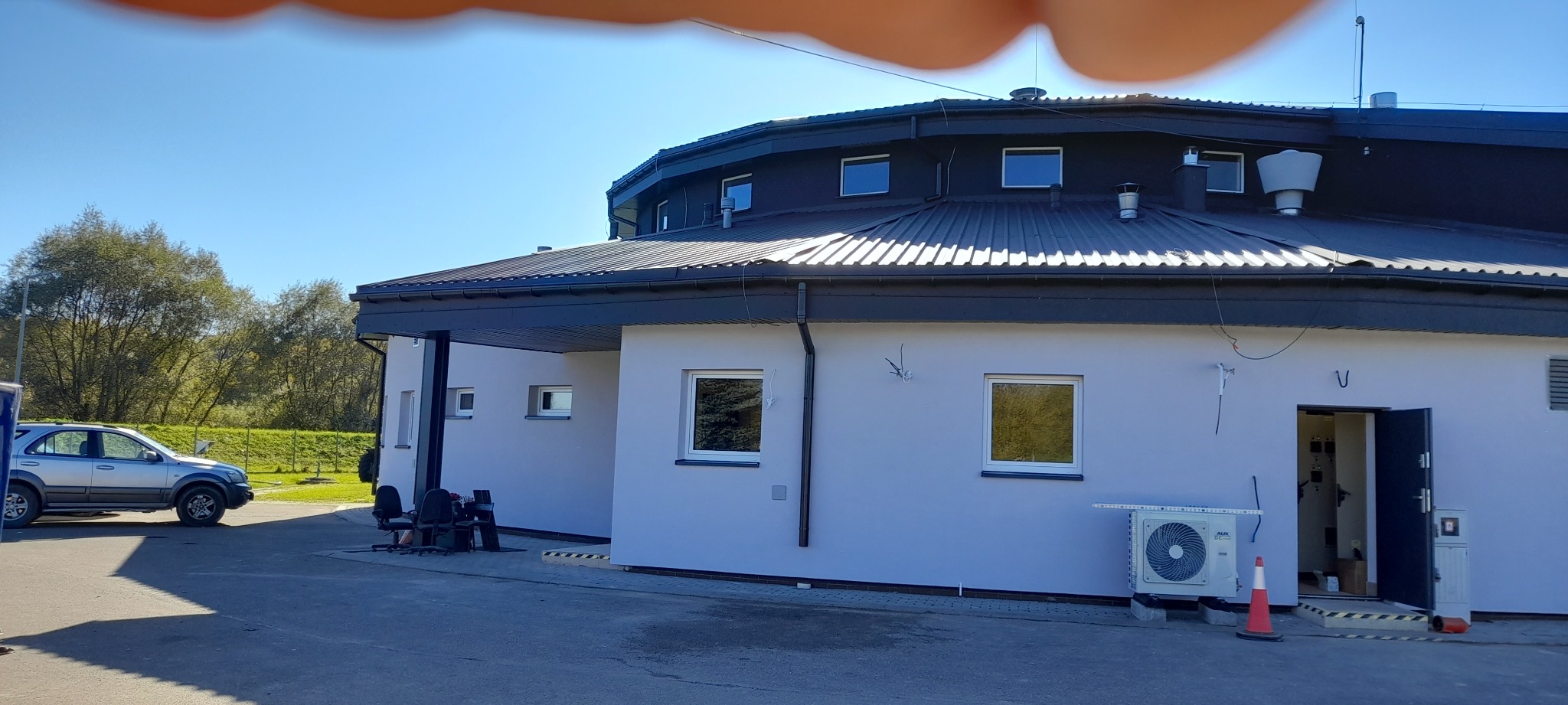 Elewacja budynku po tynkowaniu i malowaniu na szaro, z widocznym klimatyzatorem i otwartymi drzwiami. Na dachu czarna blacha. Na zewnątrz zaparkowany samochód.