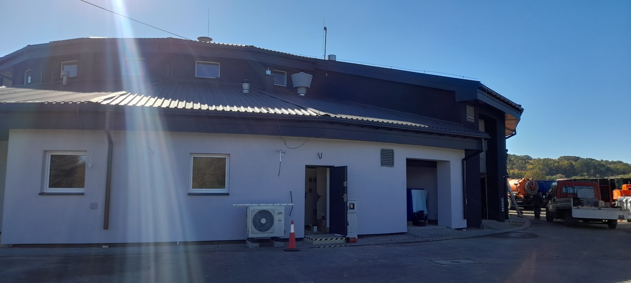 Budynek po renowacji elewacji z klimatyzatorem na zewnątrz. Widoczne okna, drzwi wejściowe i brama garażowa. W tle zaparkowane pojazdy i zieleń.