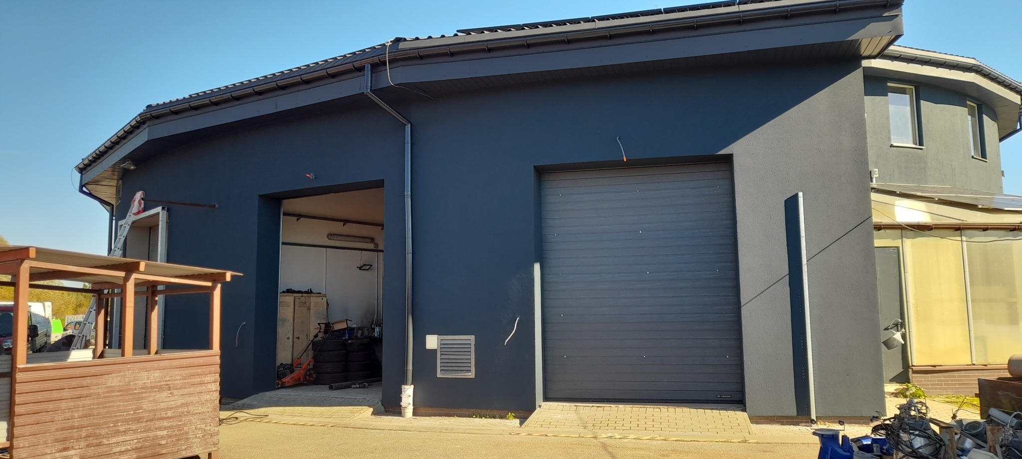 Ciemnoszara elewacja budynku z dwoma bramami garażowymi, jedna otwarta, druga zamknięta. Widoczne elementy konstrukcyjne i otoczenie budynku w słoneczny dzień.