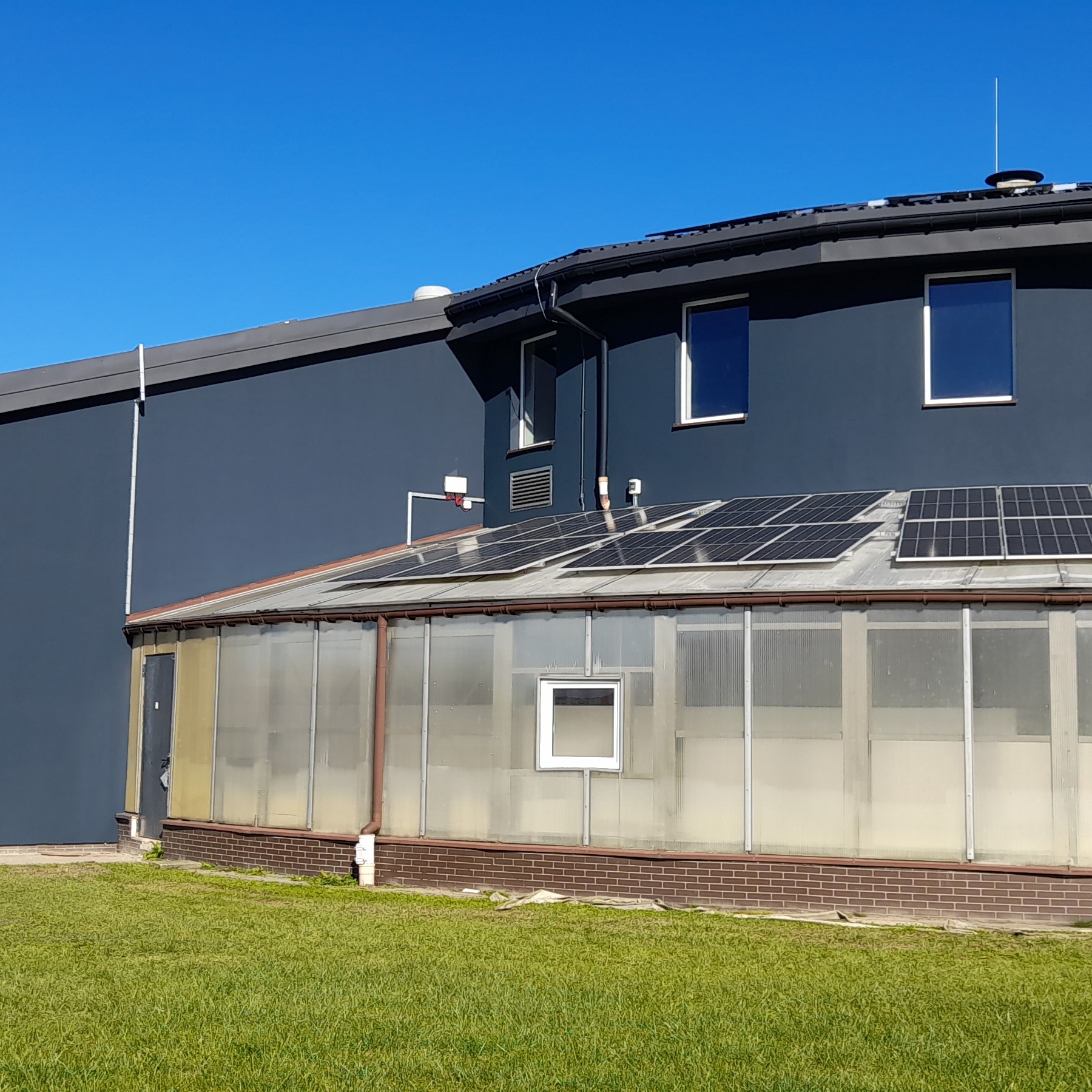 Nowoczesny budynek z ciemną elewacją i panelami słonecznymi na dachu oranżerii. Widoczne okna i fragment trawnika. Jasne, słoneczne oświetlenie.