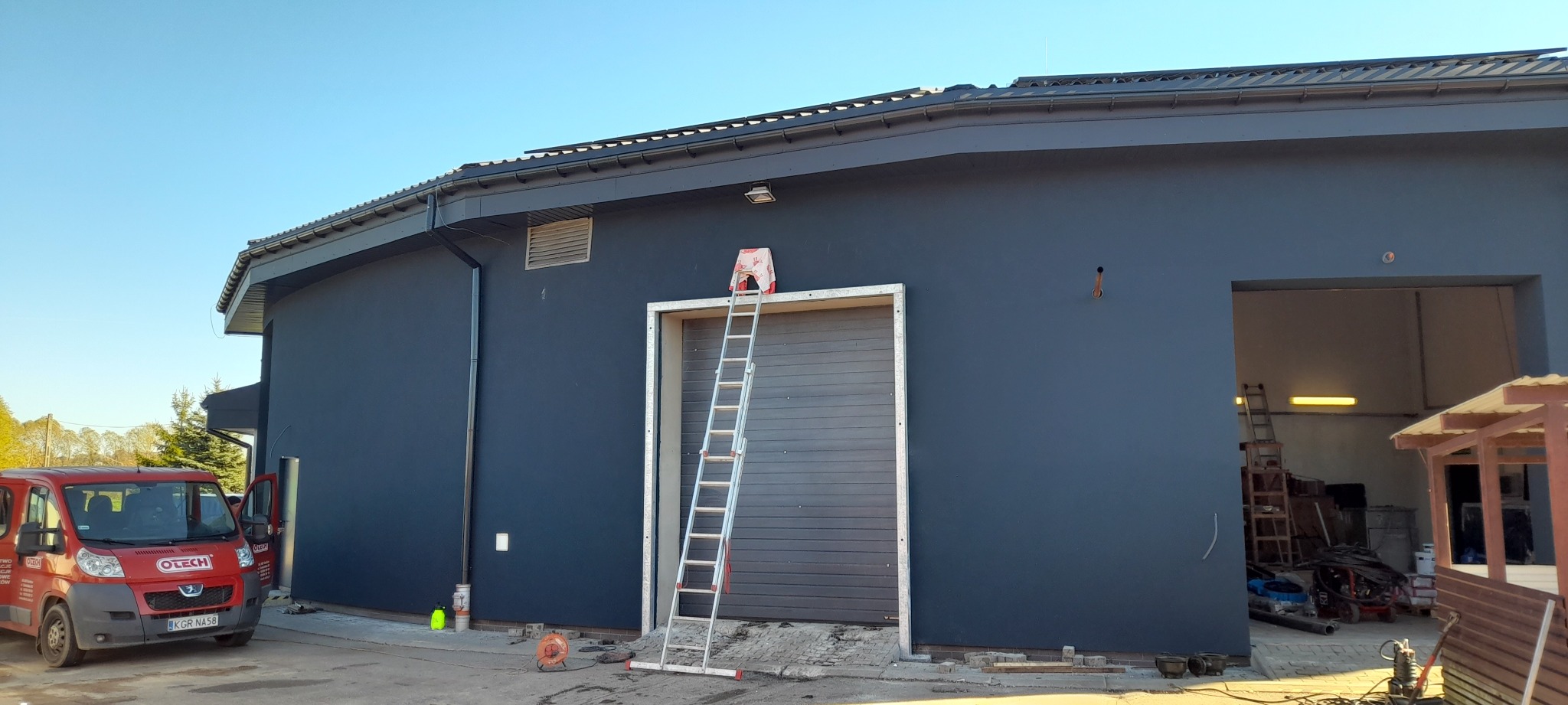Ciemnoszara elewacja budynku z drabiną opartą o bramę garażową. Widoczny samochód dostawczy firmy i narzędzia. Słoneczny dzień.