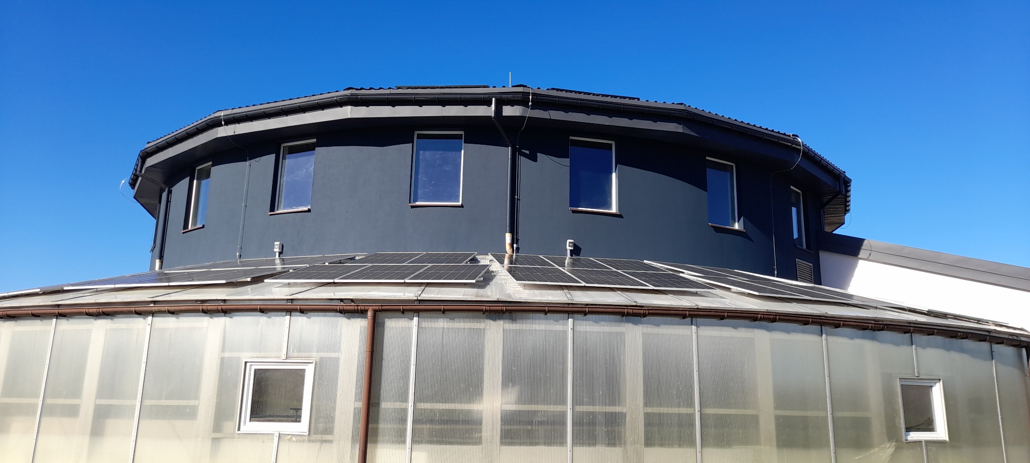 Oryginalny budynek o nietypowej architekturze z ciemną elewacją i panelami słonecznymi na dachu, widziany z dołu na tle błękitnego nieba.