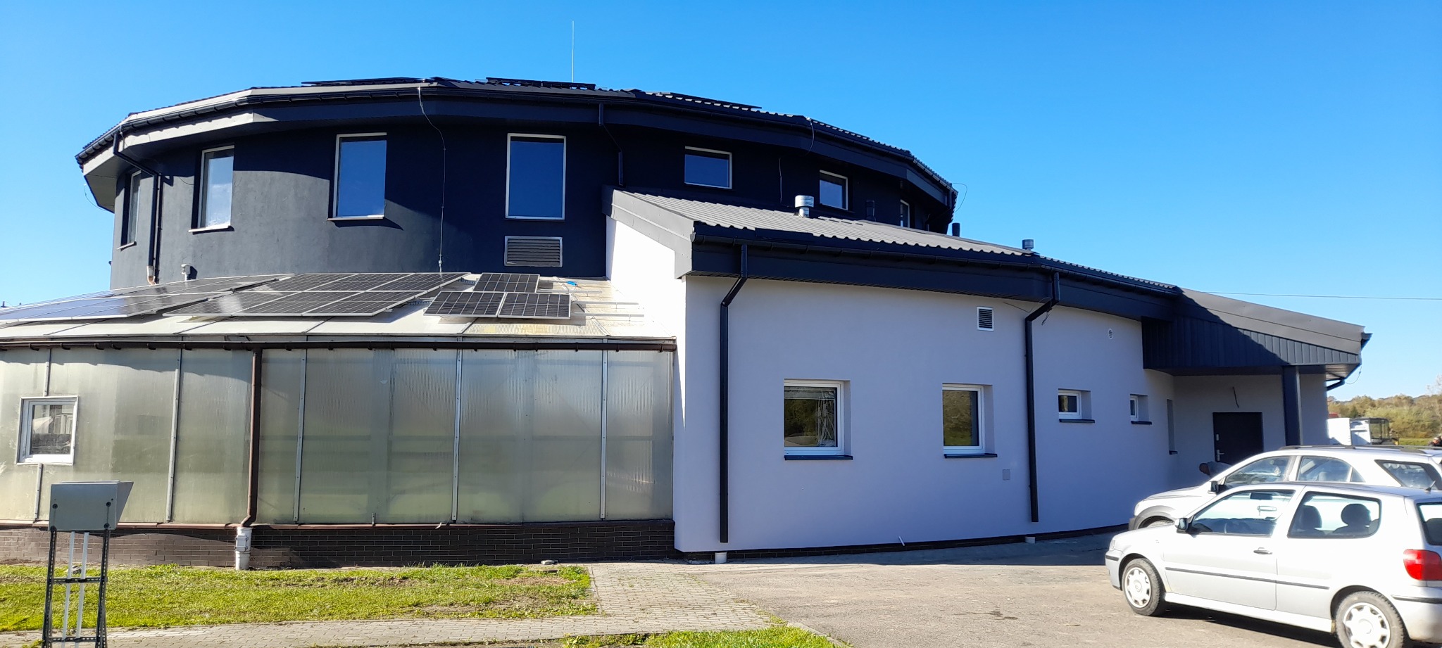 Dwukondygnacyjny budynek z panelami słonecznymi i szklarnią, pomalowany na szaro i czarno, z widocznymi samochodami na parkingu w słoneczny dzień.
