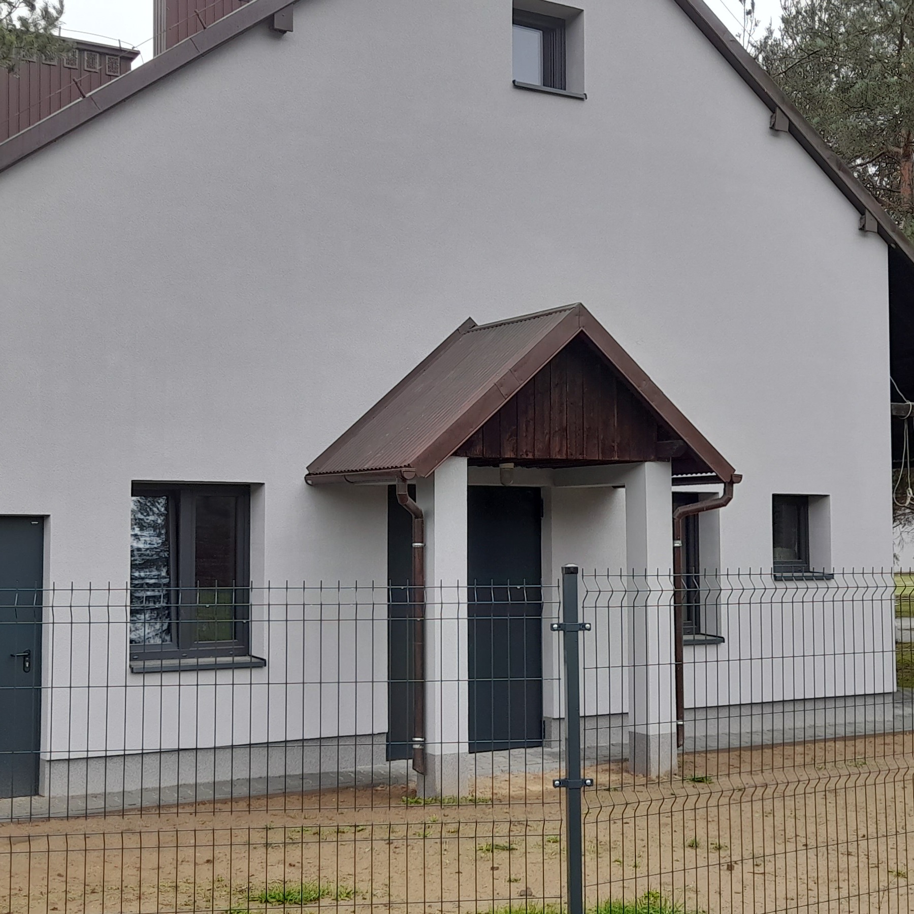 Dom jednorodzinny z jasną elewacją i ciemnobrązowym dachem, widoczny zza metalowego ogrodzenia. Detal: zadaszenie nad wejściem z drewnianym wykończeniem.