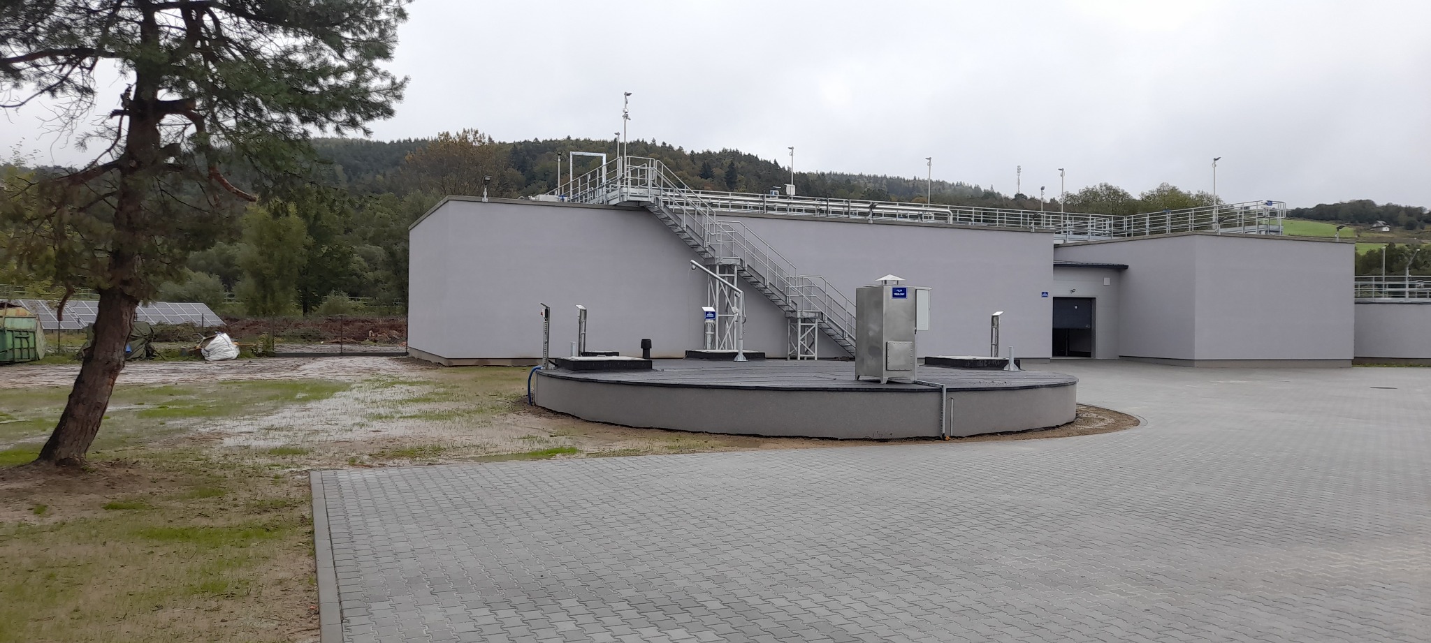 Nowoczesna oczyszczalnia ścieków z szarymi budynkami i metalowymi schodami na tle zalesionych wzgórz w pochmurny dzień. Widoczny fragment drzewa i kostka brukowa.