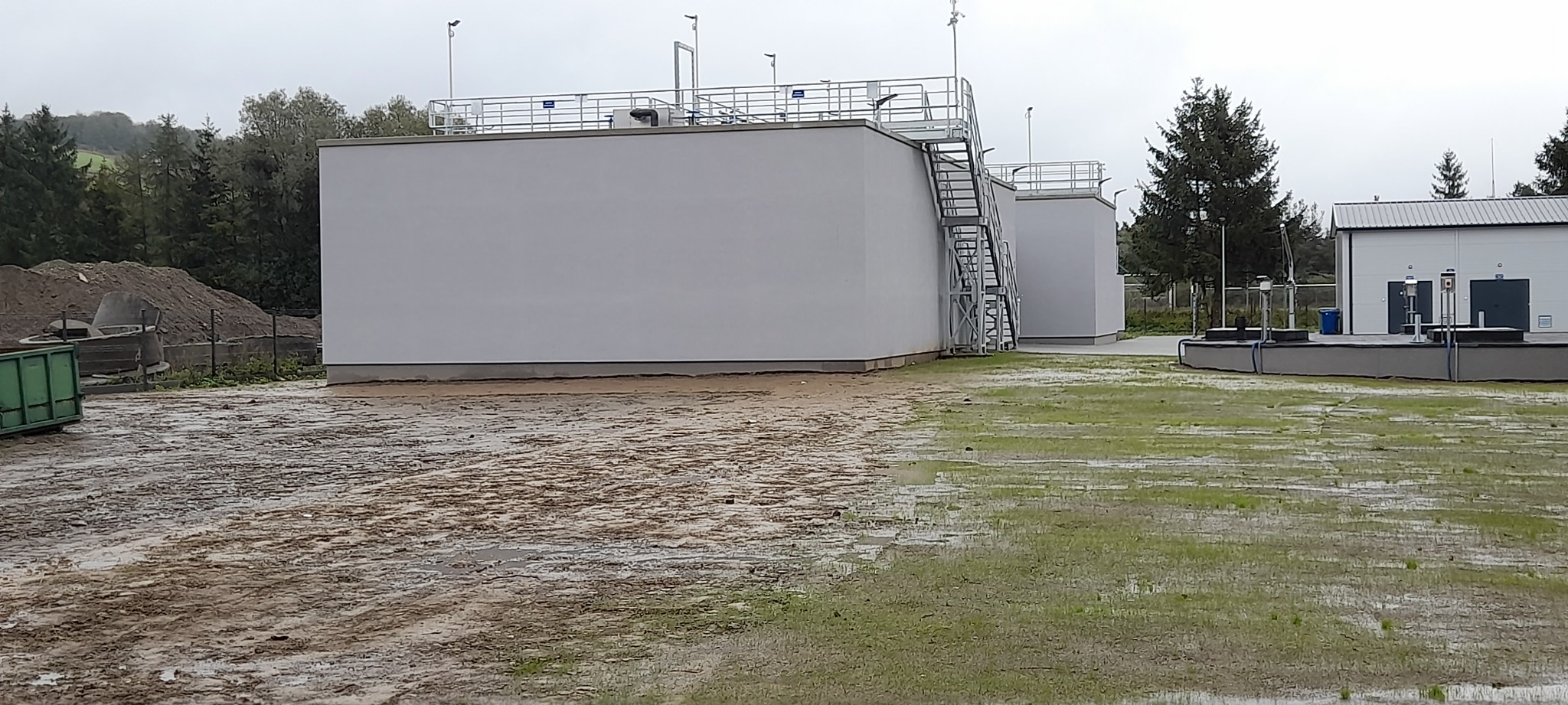 Białe, prostopadłościenne zbiorniki z metalowymi schodami na zewnątrz, otoczone mokrym terenem i zielenią, w tle drzewa i budynek. Pochmurny dzień.