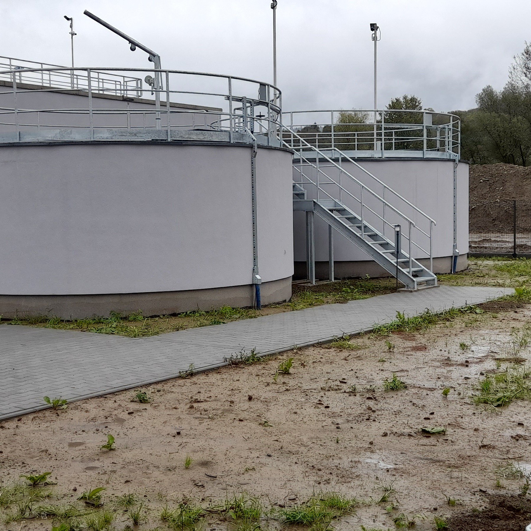 Dwa cylindryczne zbiorniki z szarą elewacją i metalowymi schodami na zewnątrz, otoczone kostką brukową i błotnistym terenem. Pochmurne niebo w tle.