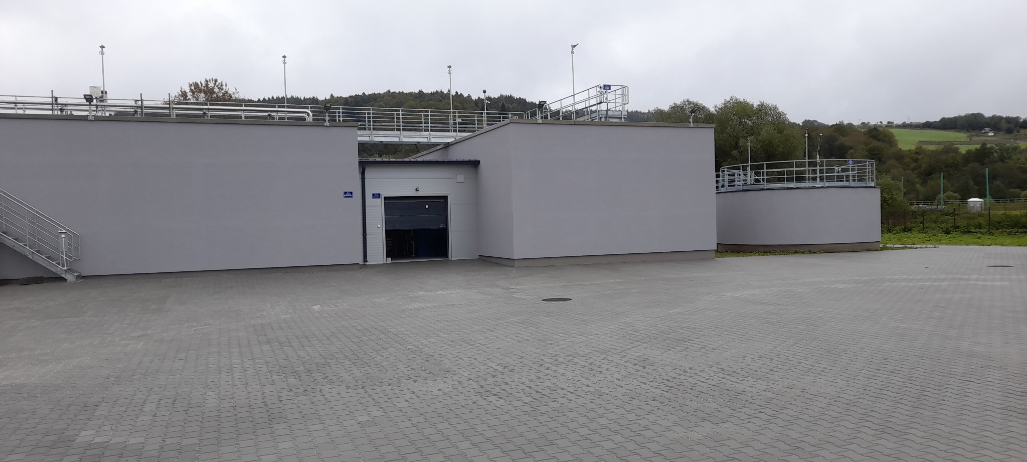 Szary budynek przemysłowy z otwartą bramą garażową i metalowymi schodami na zewnątrz, na tle pochmurnego nieba i zielonych wzgórz. Widoczna kostka brukowa.