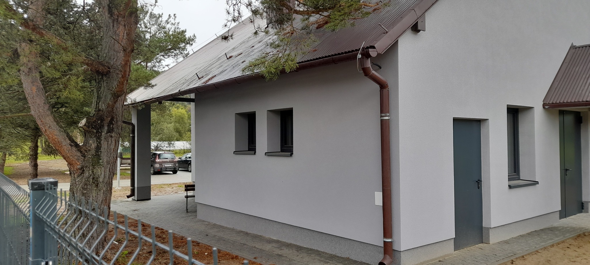 Elewacja budynku po remoncie, z widocznymi oknami, drzwiami i rynną w kolorze brązowym. Tło stanowią drzewa i częściowo widoczny samochód.