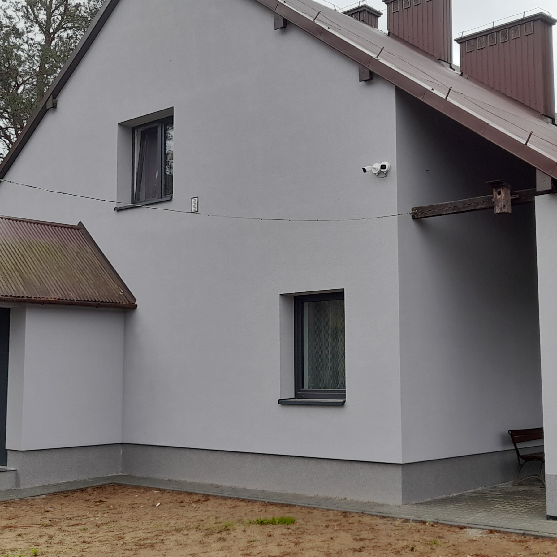 Elewacja domu w odcieniach szarości z brązowym dachem, widoczne okna z ciemnymi ramami, kamera monitoringu i fragment drewnianej konstrukcji. Ujęcie z zewnątrz.