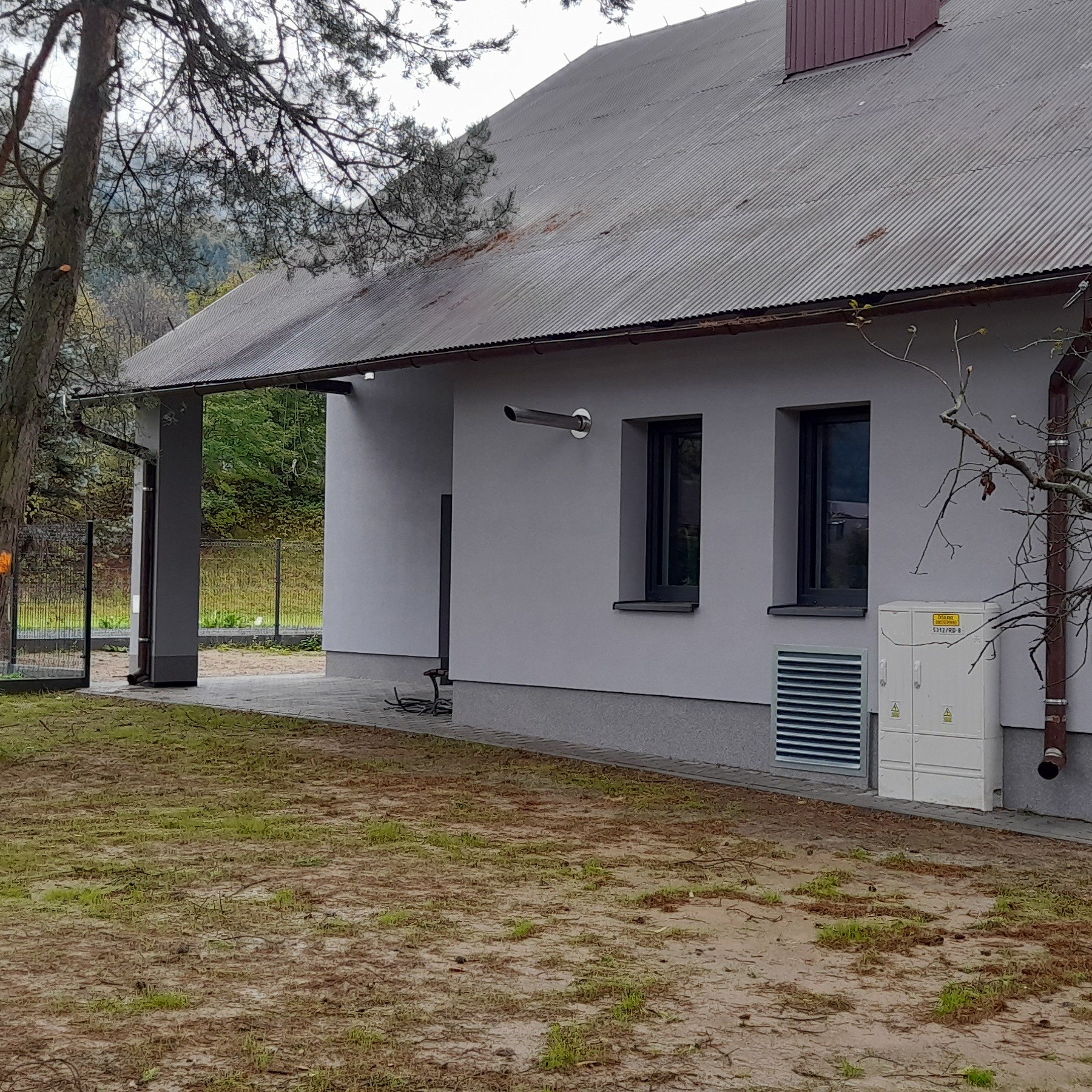 Jasnoszara elewacja domu z ciemnymi oknami, widoczny fragment dachu z blachy falistej, skrzynka elektryczna i trawnik z rzadką trawą. Ujęcie z boku budynku.