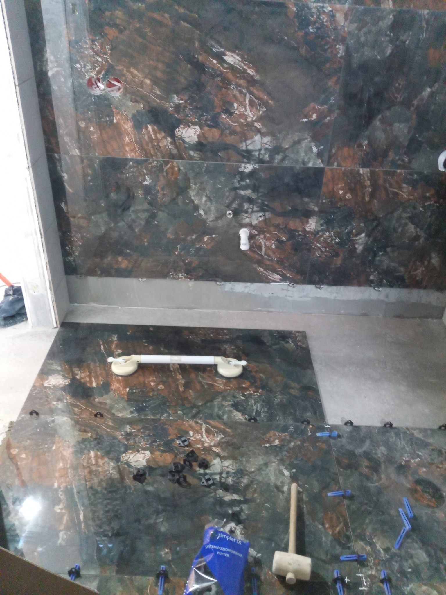 Układanie płytek imitujących marmur w łazience, widoczne przyssawki do przenoszenia płytek, młotek gumowy i system poziomowania płytek.