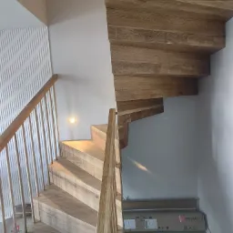 Drewniane schody spiralne z jasnego drewna, z minimalistyczną balustradą, oświetleniem punktowym na ścianie i dekoracyjną ścianą z pionowymi panelami, pudełko z listwami przypodłogowymi na podłodze.