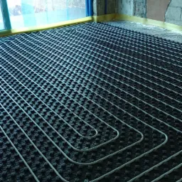 Rozłożona instalacja ogrzewania podłogowego na czarnej macie izolacyjnej w surowym pomieszczeniu z oknem zabezpieczonym folią malarską.