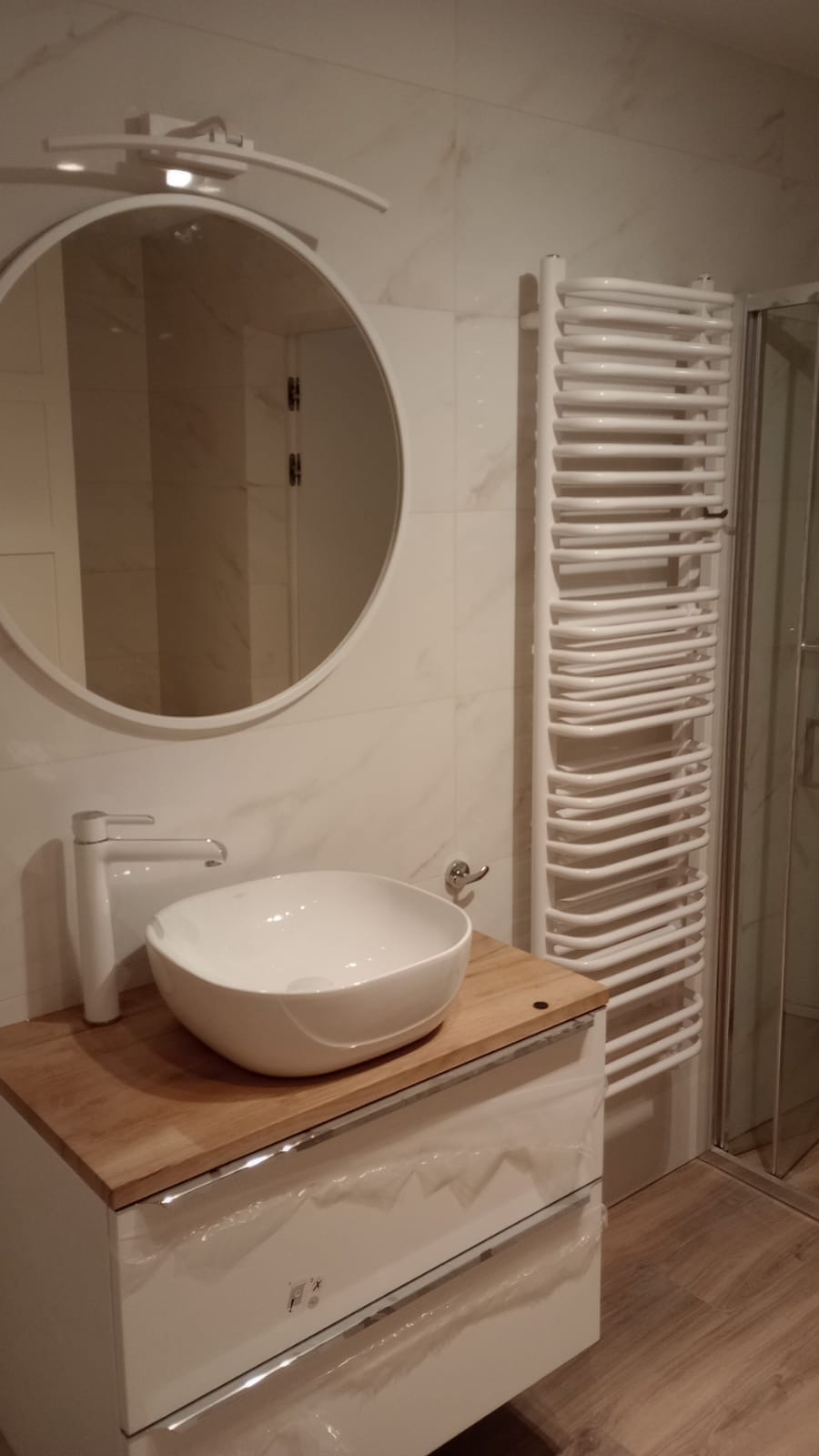 Biała umywalka nablatowa na drewnianym blacie z szafką, okrągłe lustro z oświetleniem, biały grzejnik drabinkowy i kabina prysznicowa w tle, marmurowe płytki na ścianie.