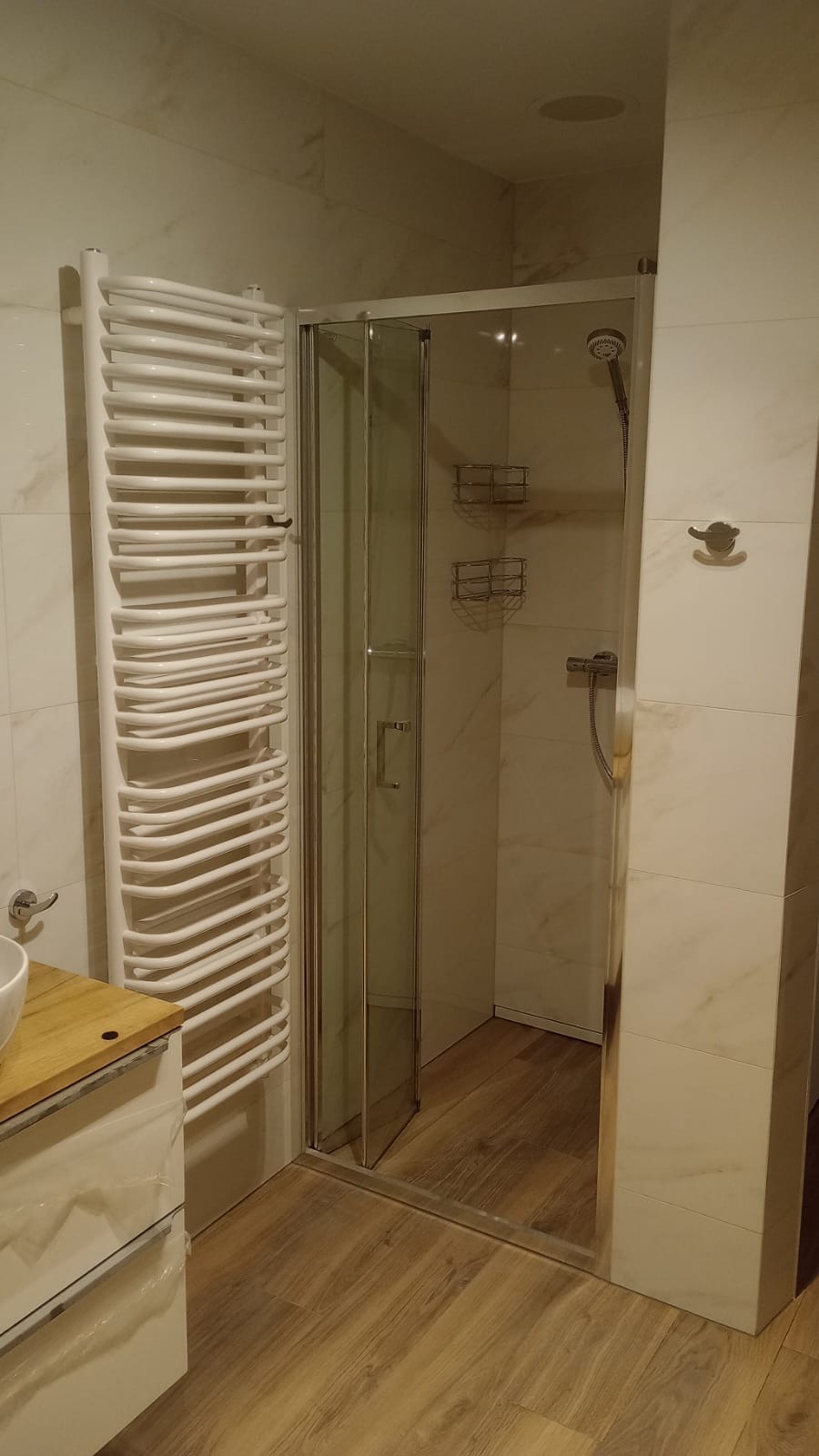 Nowoczesna łazienka z kabiną prysznicową z przezroczystego szkła, grzejnikiem drabinkowym w kolorze białym, marmurowymi płytkami ściennymi i podłogą imitującą drewno.