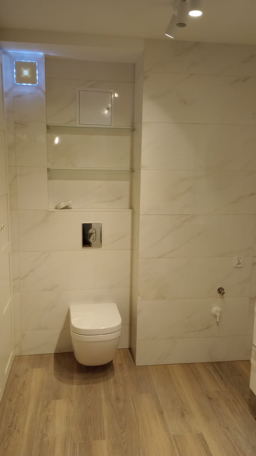 Nowoczesna toaleta podwieszana w łazience wykończonej białymi płytkami imitującymi marmur i drewnopodobną podłogą, z półkami szklanymi i oświetleniem punktowym.