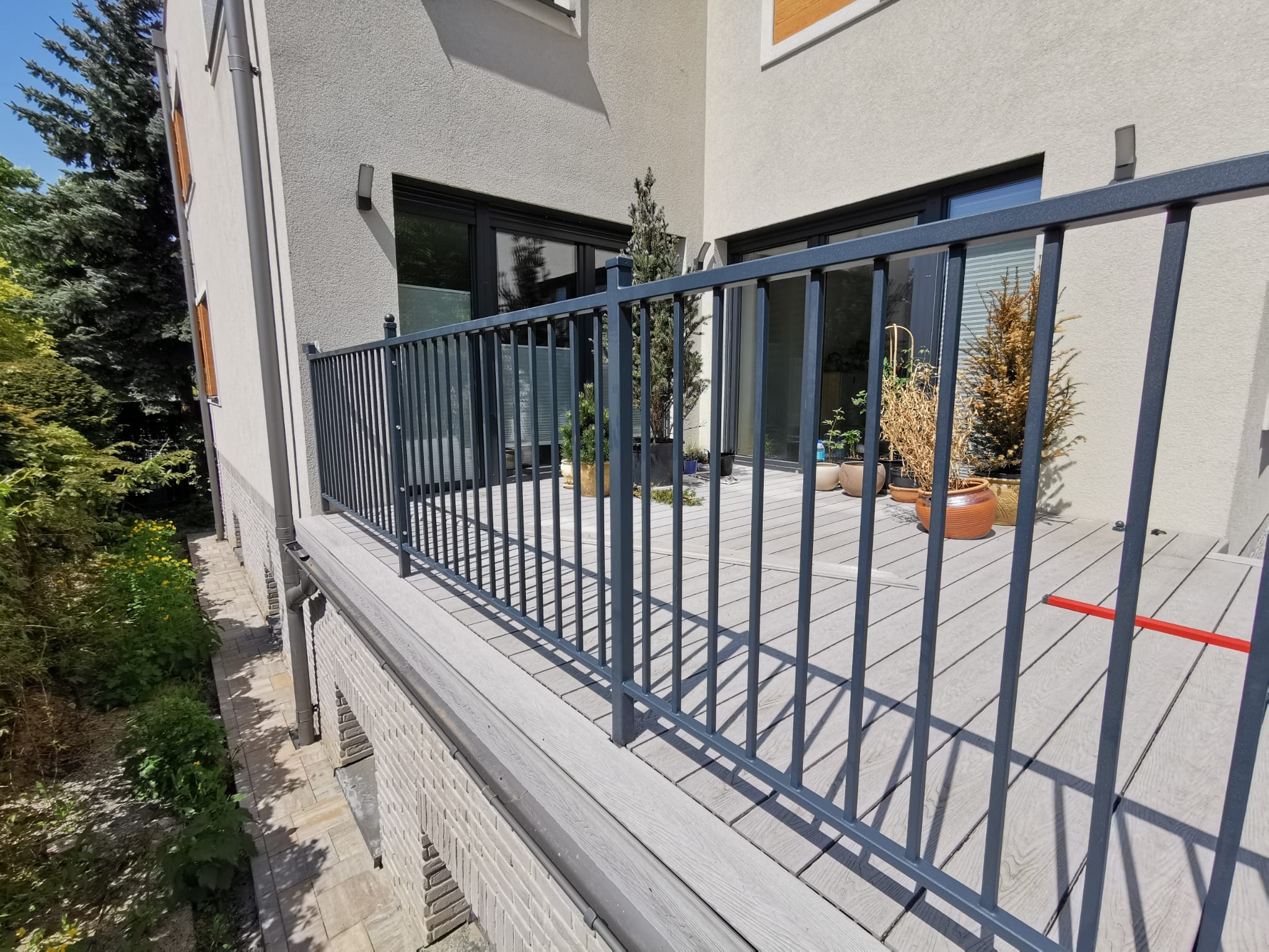 Nowoczesny balkon z szarą balustradą i kompozytową podłogą, widok z zewnątrz na rośliny w donicach i elewację budynku.