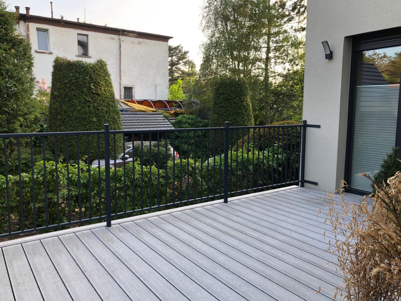 Nowoczesny taras z szarą deską kompozytową i czarną metalową balustradą we Wrocławiu, widok na zielony ogród i budynek w tle.