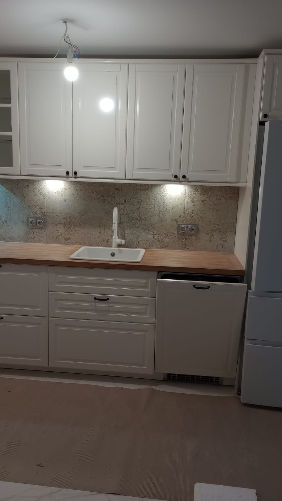 Biała zabudowa kuchenna z drewnianym blatem i szarą mozaiką na ścianie, oświetlona punktowymi lampami podszafkowymi, z widoczną instalacją elektryczną oświetlenia górnego.