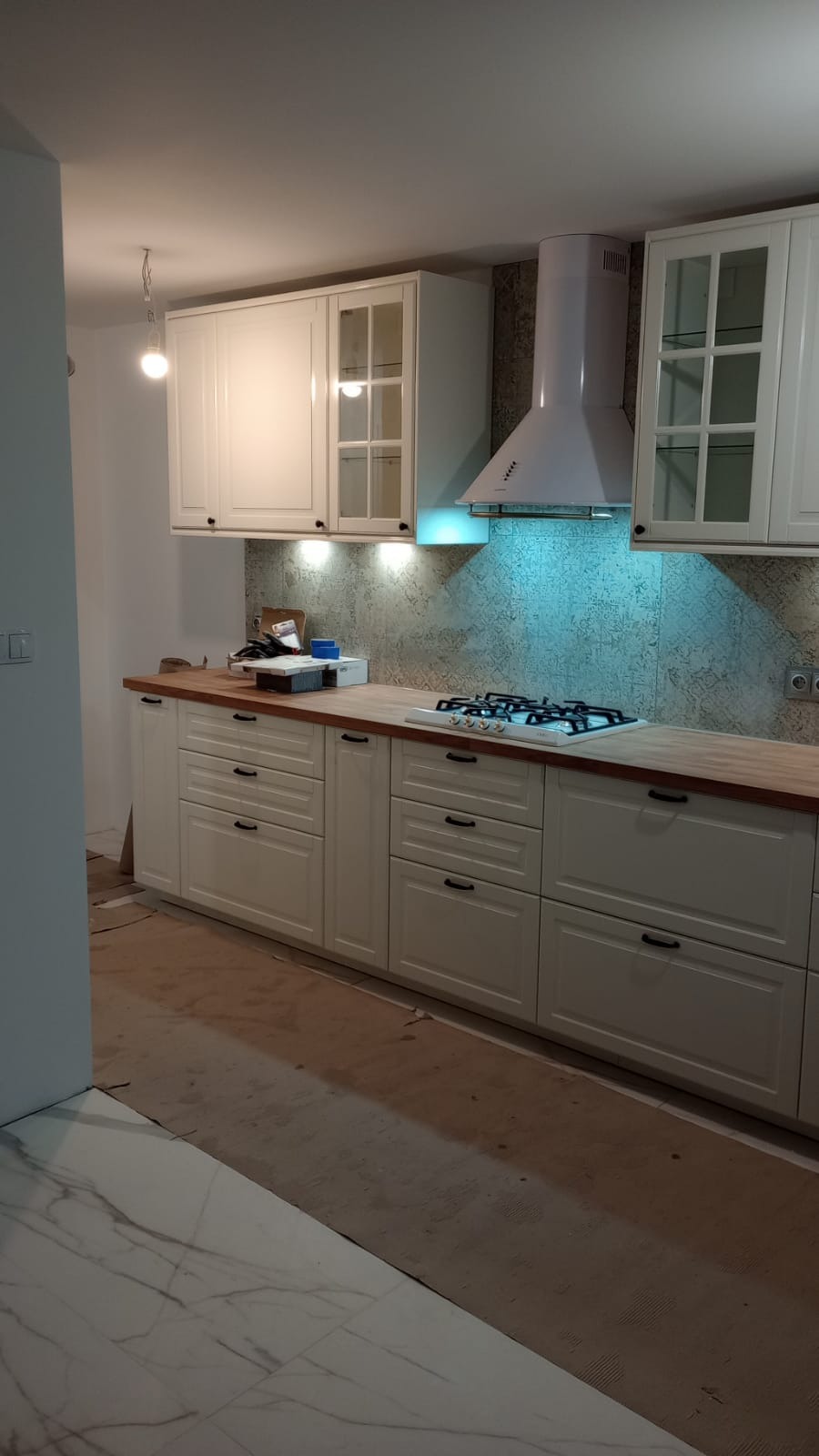 Montaż białej zabudowy kuchennej z drewnianym blatem, widoczny fragment nowej marmurowej podłogi, okap i płyta gazowa.