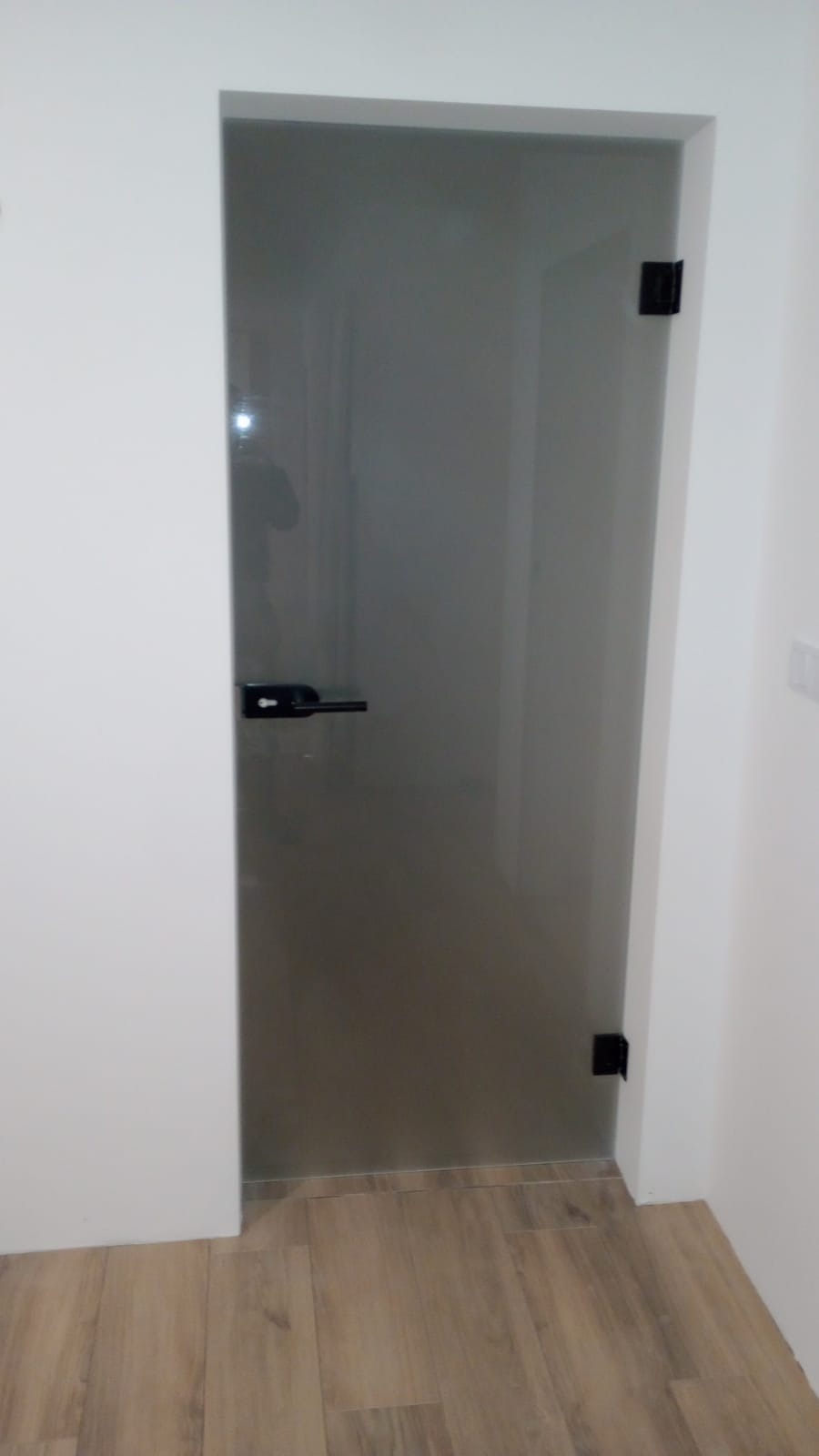 Szklane drzwi z czarną klamką i zawiasami, zamontowane w białej ścianie, z widoczną podłogą imitującą drewno.