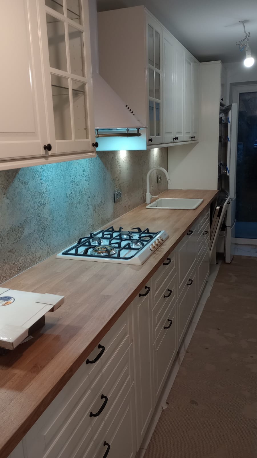 Nowoczesna kuchnia z białymi szafkami z frezowanymi frontami i drewnianym blatem, płyta gazowa z białym obramowaniem, biały zlew i bateria, szary wzorzysty panel nad blatem, oświetlenie podszafkowe.