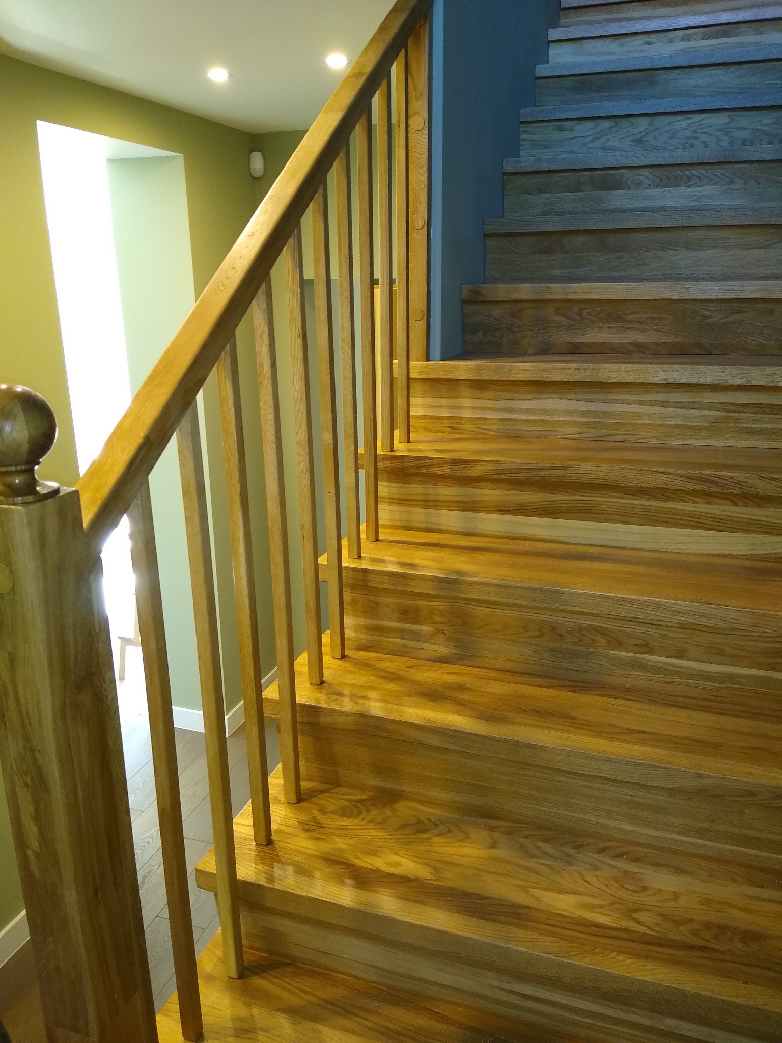 Drewniane schody z jasną balustradą w nowoczesnym wnętrzu, widoczne różne odcienie drewna i oświetlenie punktowe.