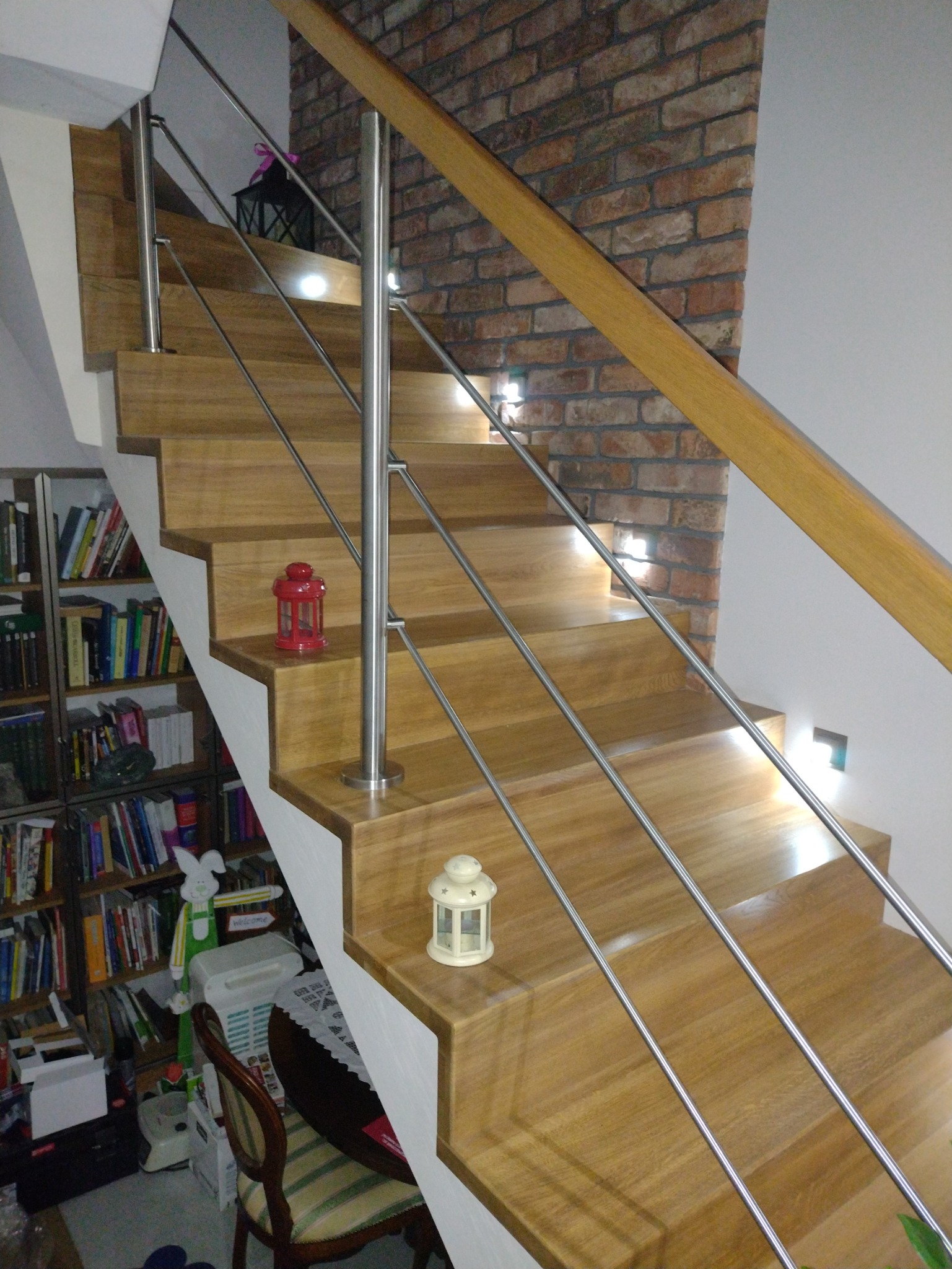 Drewniane schody z metalową balustradą, oświetleniem punktowym i dekoracjami w postaci lampionów, na tle ściany z cegły i regału z książkami.