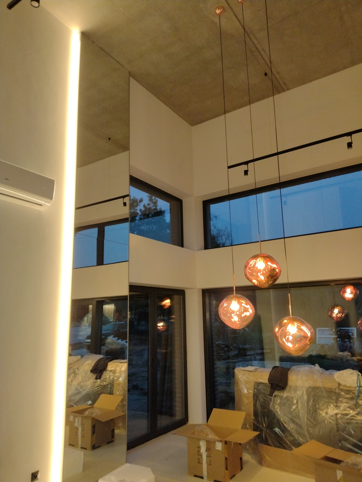 Wnętrze w trakcie wykańczania: surowy betonowy sufit, nowoczesne lampy wiszące z miedzianego szkła, duże okna z widokiem na zewnątrz, kartony i folia ochronna na podłodze, pionowe oświetlenie LED...