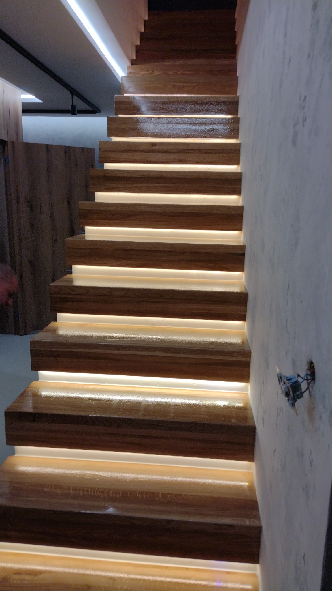 Drewniane schody z podświetleniem LED między stopniami, widok z dołu, surowa ściana obok z wystającymi przewodami elektrycznymi.