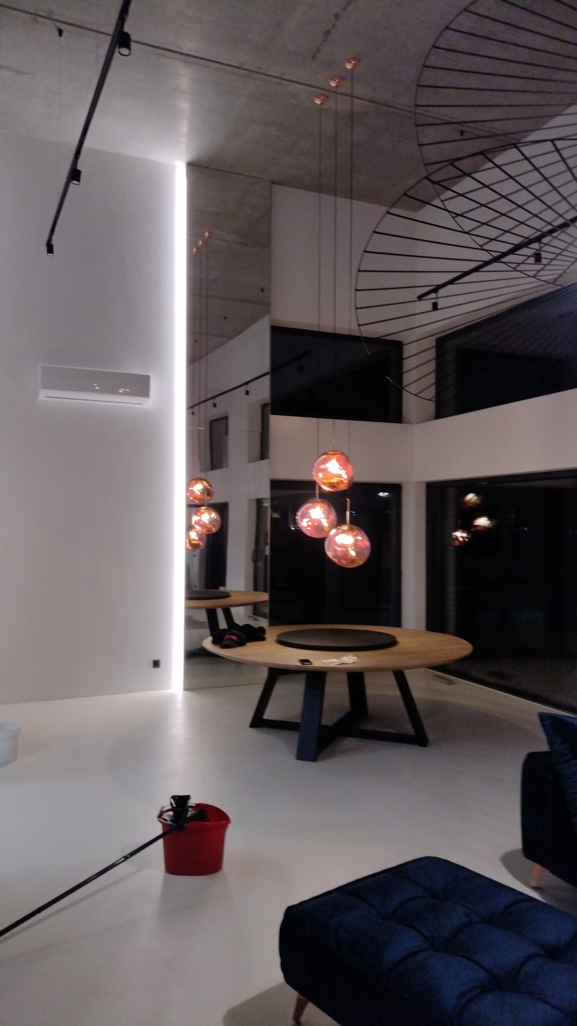 Nowoczesne wnętrze z okrągłymi drewnianymi stolikami, wiszącymi lampami w kształcie kul oraz wiadrem z mopem na białej podłodze.