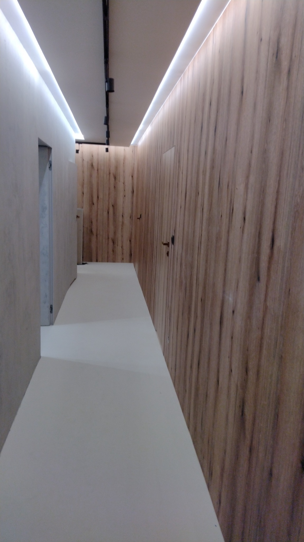 Nowoczesny korytarz z podświetleniem LED na suficie i ścianami wykończonymi panelami imitującymi drewno, widoczne drzwi po obu stronach.