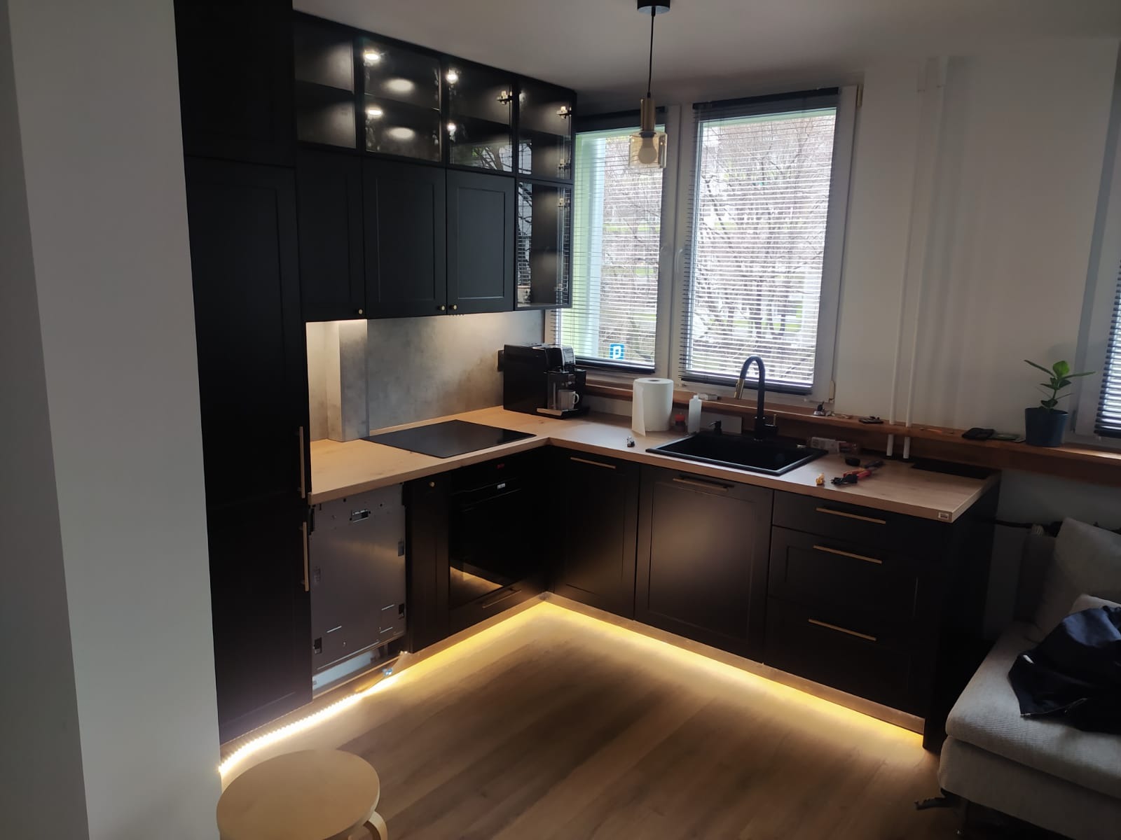 Czarna, narożna zabudowa kuchenna z drewnianym blatem i podświetleniem LED przy podłodze oraz szafkami wiszącymi z podświetlanymi witrynami; widoczny fragment niedokończonego montażu z otwartą...