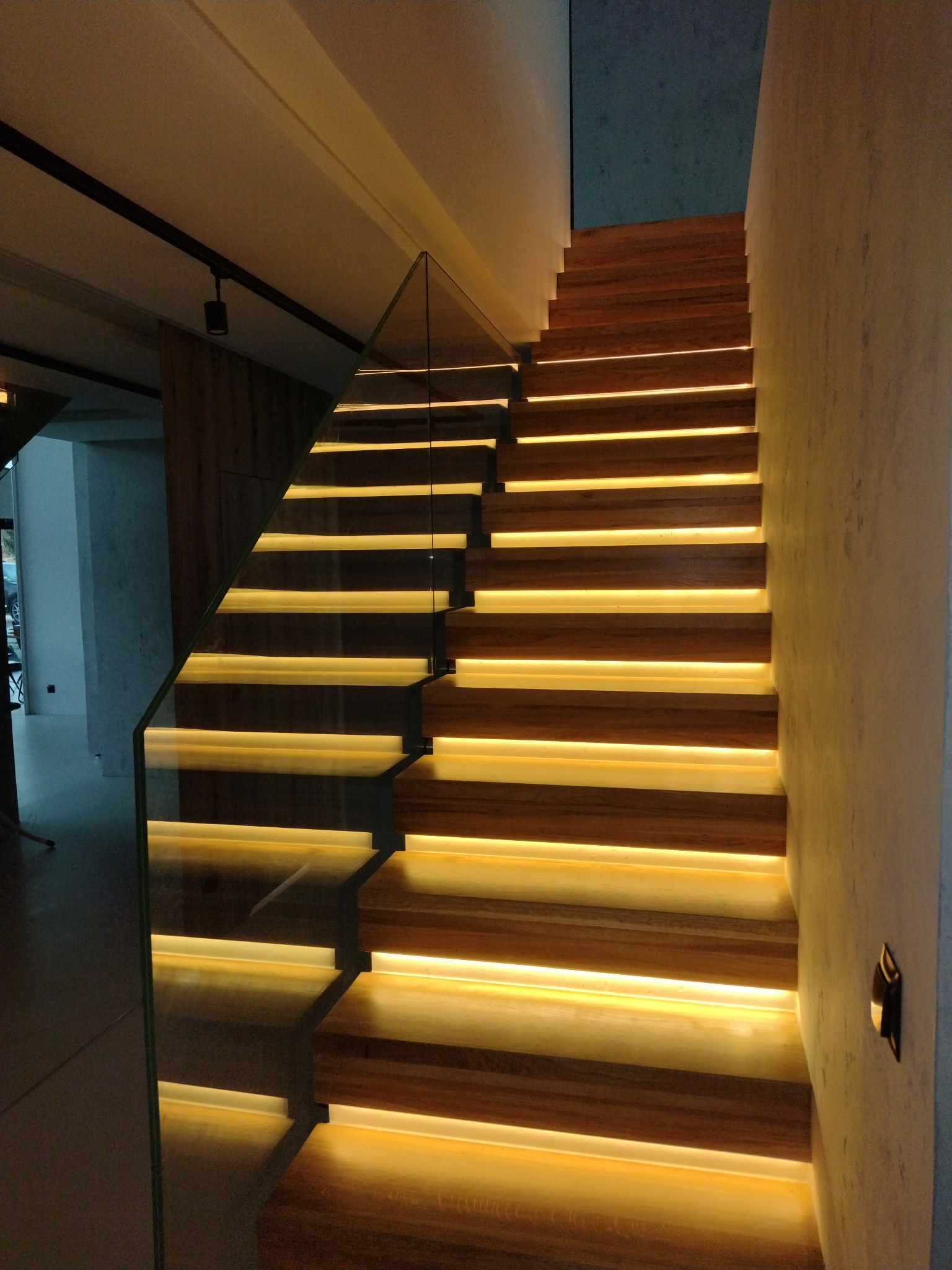 Nowoczesne drewniane schody z podświetleniem LED na każdym stopniu i szklaną balustradą, oświetlenie punktowe na suficie.