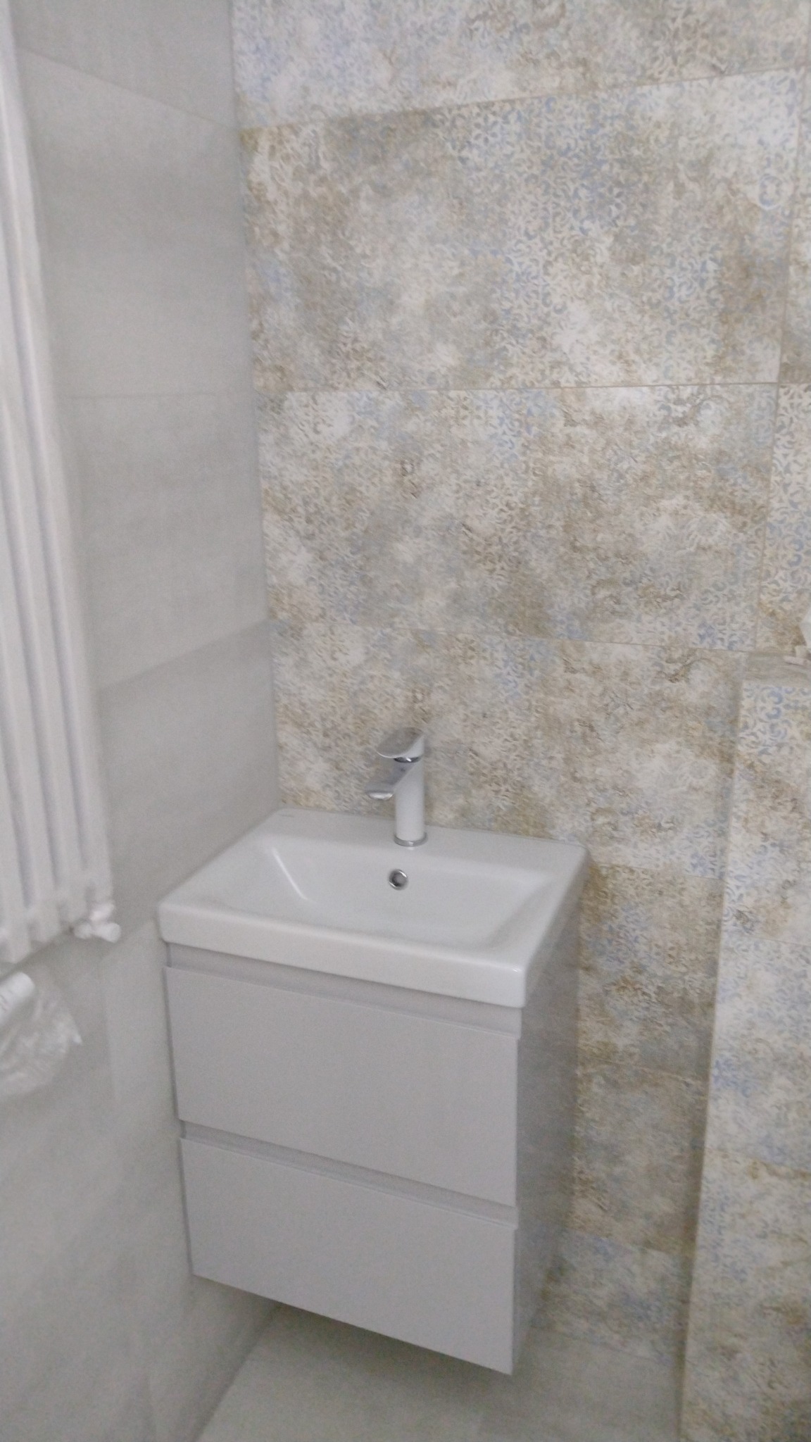 Biała umywalka z szafką w łazience wykończonej płytkami w odcieniach beżu i błękitu, widoczny chromowany kran.