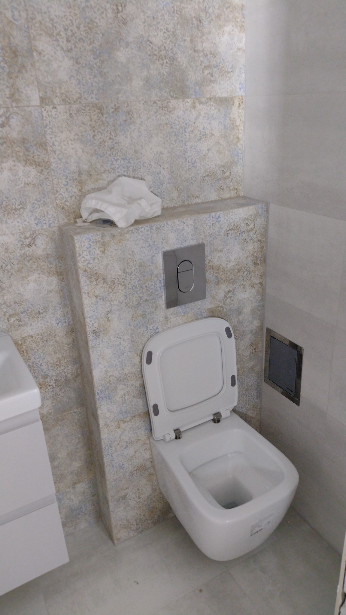 Nowoczesna, podwieszana toaleta z otwartą klapą, zamontowana na ścianie wykończonej dekoracyjnymi płytkami w odcieniach beżu i błękitu, z widocznym przyciskiem spłukiwania i fragmentem umywalki po...