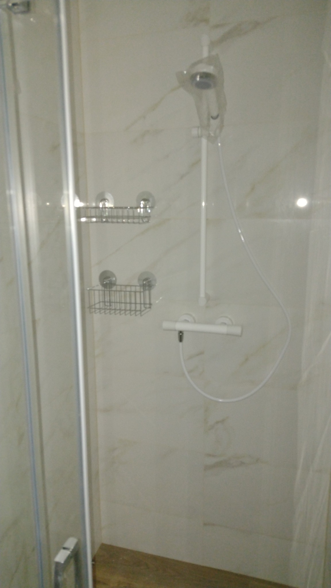 Nowa kabina prysznicowa z marmurowymi płytkami, deszczownicą w folii ochronnej i metalowymi półkami na przyssawki.