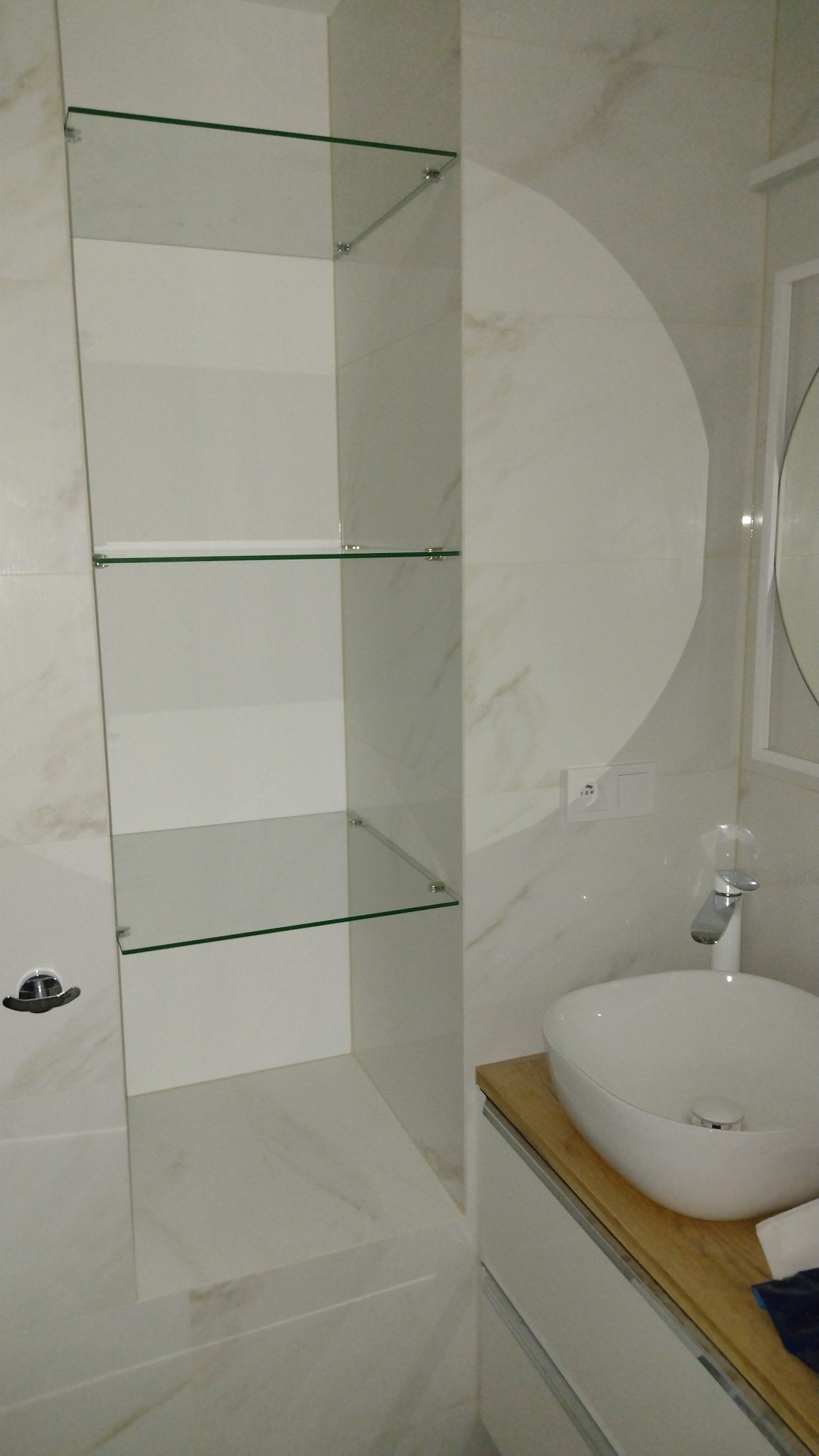 Nowoczesna łazienka z umywalką nablatową, baterią i szklanymi półkami w zabudowie, wykończona białymi płytkami imitującymi marmur.