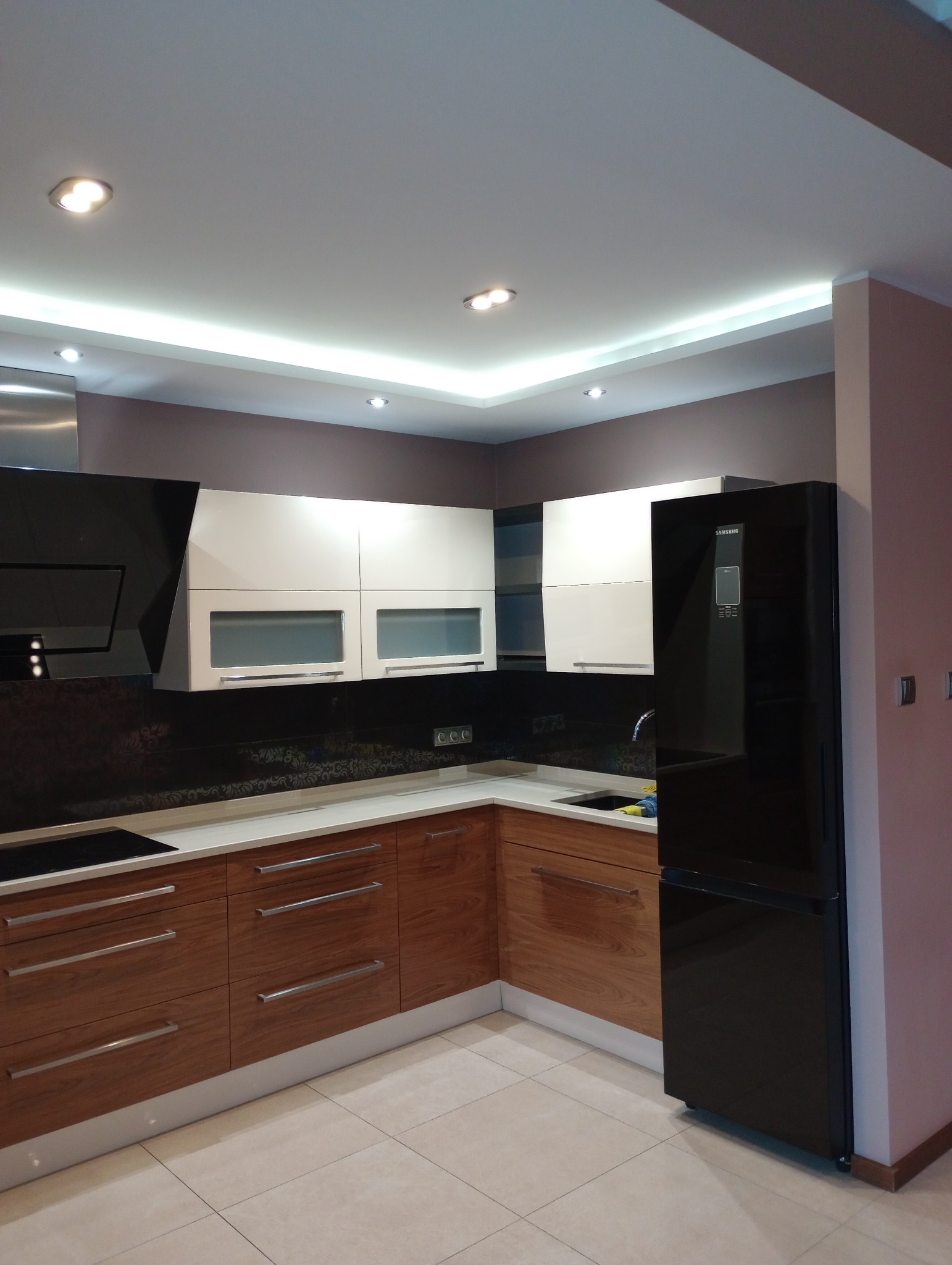 Nowoczesna kuchnia z drewnianymi szafkami dolnymi, białymi szafkami górnymi z przeszkleniami, czarnym panelem nad blatem i czarną lodówką Samsung, oświetlona ledowym paskiem pod sufitem