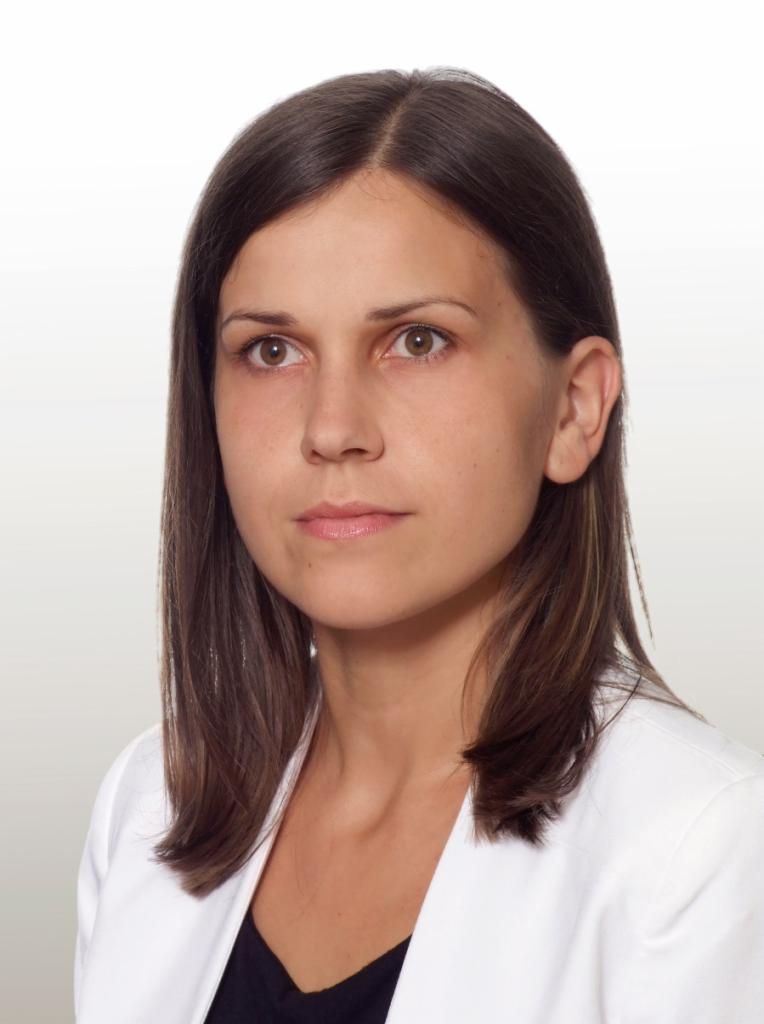 Portret kobiety w białym żakiecie na jasnym tle, patrzącej w bok z lekkim uśmiechem.