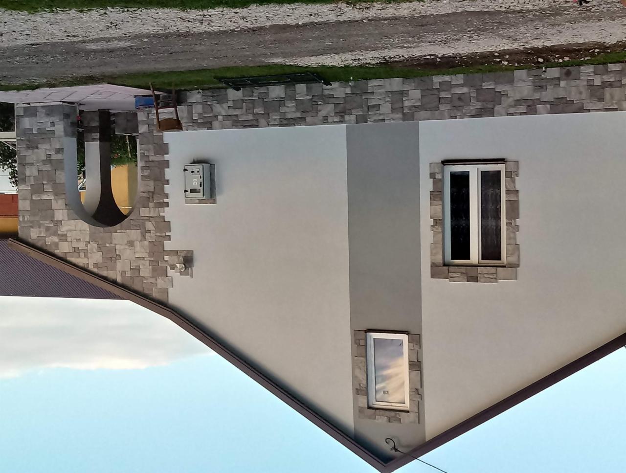 Jasnoszara elewacja domu jednorodzinnego z dekoracyjnym obramowaniem okien i łukowym wejściem wykończonym kamieniem elewacyjnym.