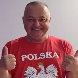 Uśmiechnięty mężczyzna w czerwonej koszulce z napisem POLSKA i orłem w koronie, pokazujący kciuki w górę na tle jasnej ściany.