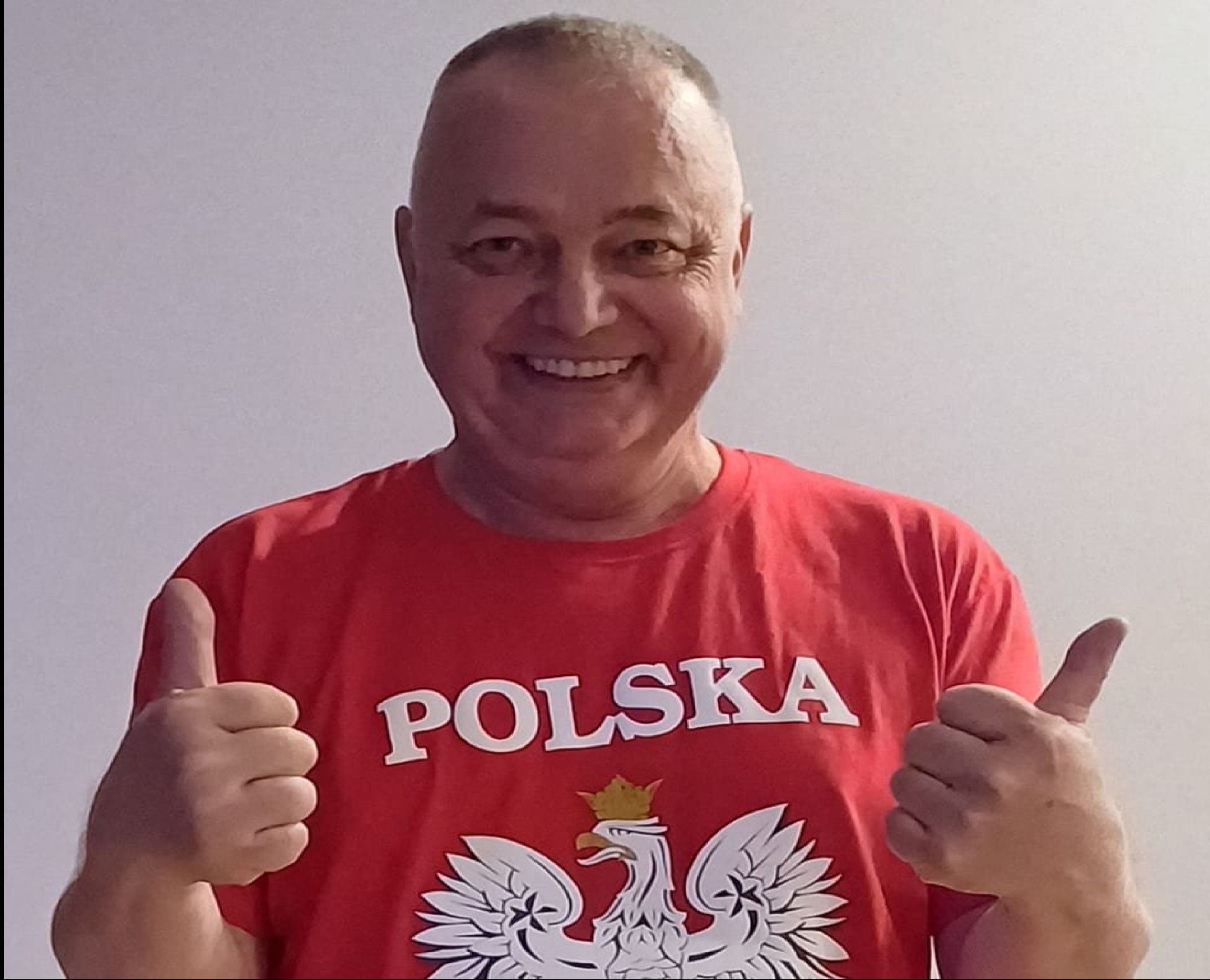 Uśmiechnięty mężczyzna w czerwonej koszulce z napisem POLSKA i orłem w koronie, pokazujący kciuki w górę na tle jasnej ściany.