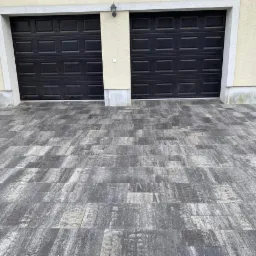 Ułożona kostka brukowa w odcieniach szarości i bieli przed garażem z dwiema czarnymi bramami segmentowymi, widoczne delikatne różnice w odcieniach kostki.
